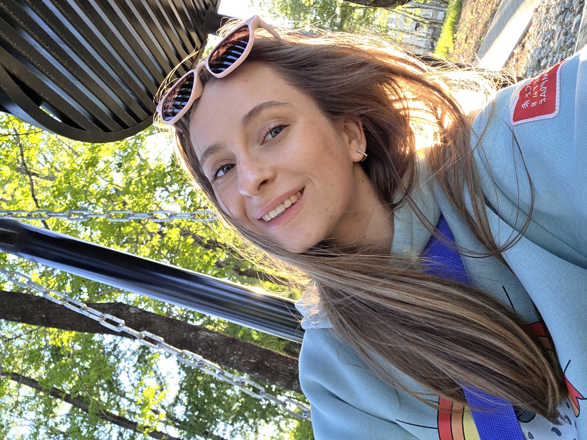 A fair-skinned woman with long brown hair and pink sunglasses perched on her head smiles at the camera. She wears a light blue hoodie with a red patch on the upper left sleeve, and a blue lanyard around her neck. The shot is taken from below, with the woman’s face dominating the frame. Behind her, a structure of black metal bars and chains is partially visible, with green trees and foliage forming a backdrop. The light suggests it is daytime, and the presence of trees indicates an outdoor setting.
The city Birobidzhan and country Russia are not discernable from the image.