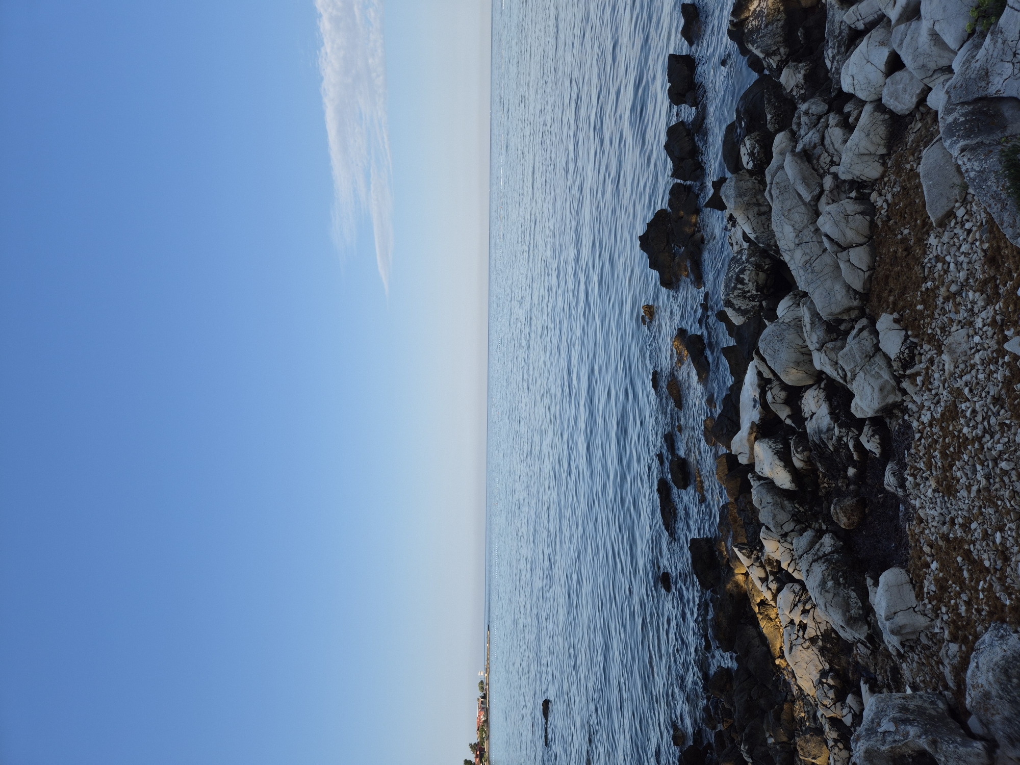The image depicts a serene coastal scene with a clear blue sky and calm sea. The foreground is dominated by a rocky shoreline, with a variety of stones and pebbles. Some of these rocks are large and irregular, while others are smaller and more rounded. In the background, a distant coastline is visible with a few buildings and trees. The water is a deep blue, reflecting the sky, and gentle ripples are visible on its surface. A thin wisp of cloud stretches across the sky. The lighting suggests it is either early morning or late afternoon. The scene is static and peaceful, with no visible people or animals. Based on the style of buildings and the presence of a coastline, the location is likely a coastal area in Monterol, Croatia.