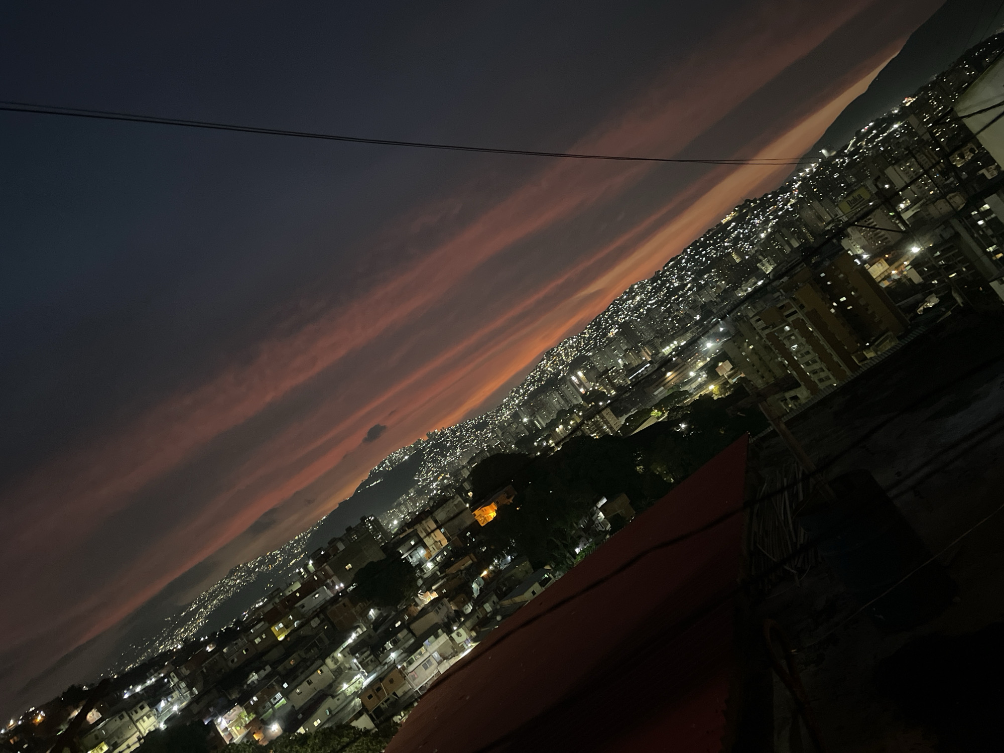 The image captures a nighttime view of Caracas, Venezuela, with a dramatic sky featuring long, reddish-pink clouds streaking across the upper portion of the frame. Below, a dense urban landscape unfolds, illuminated by countless lights from buildings and streets stretching up the mountainside. The perspective is from an elevated position, possibly a rooftop or high-rise balcony. In the foreground, a portion of a red, slightly out-of-focus rooftop is visible. Power lines cut across the upper portion of the frame, adding to the urban feel. The city appears densely populated, with closely packed buildings and a network of illuminated roadways.