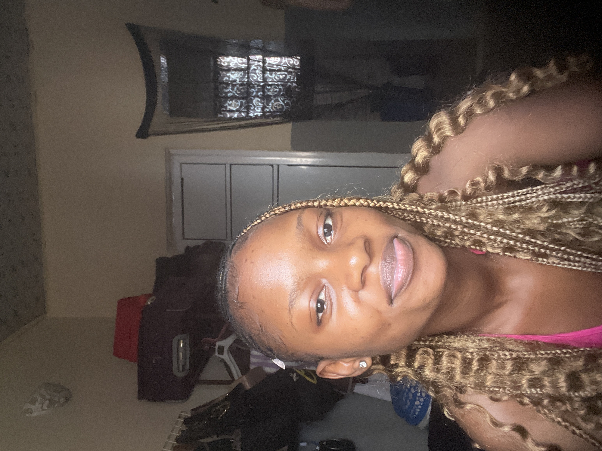 A young Black woman with light brown braids adorned with blonde highlights is shown in a close-up selfie. She is wearing a bright pink tank top. Her lips are slightly parted, and her tongue is peeking out. Her left eye is partially obscured by her hair, and she is wearing a small stud earring in her left ear. The background appears to be an indoor setting, possibly a bedroom, with a window covered by a dark, patterned curtain on the right. To the left, there are shelves with various items, including a red object on top of a dark suitcase. The lighting is dim, casting shadows on the walls and ceiling. The overall ambiance is casual and personal. This image was taken in Lafia, Nigeria.
