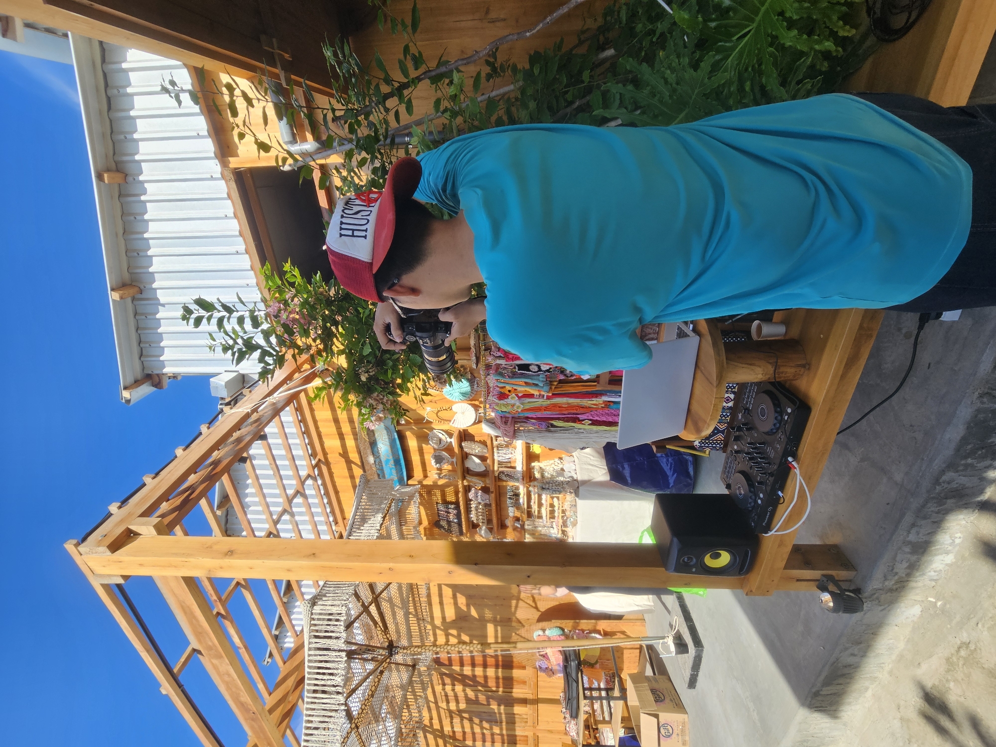 A person wearing a turquoise t-shirt and a red and white cap is taking a photo with a camera. They are standing in front of a wooden structure with a roof that appears to be under construction or renovation. To their left, a DJ setup with a laptop, mixer, and speaker is placed on a wooden table. In the background, racks of clothing and accessories are displayed, suggesting a market or retail area. The scene is outdoors, with a clear blue sky overhead and greenery visible. The location is Mũi Né, Vietnam, a popular coastal town known for its sand dunes and beaches.
