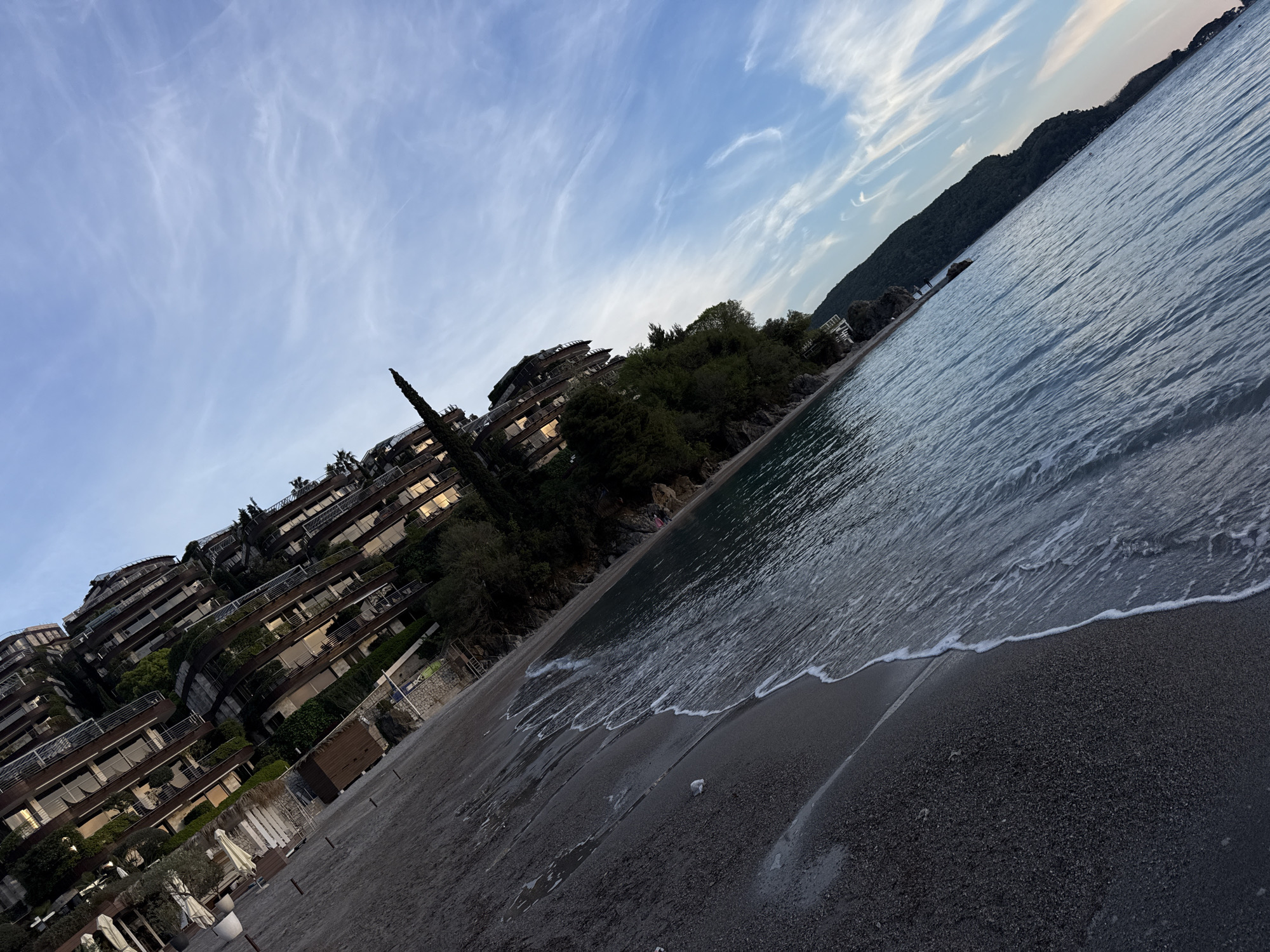 A landscape photograph of a beach on a coastline with buildings and trees. The beach is black sand with waves gently washing ashore. The water is blue-grey, and the sky is light blue with streaks of white clouds. In the distance, a mountain range is visible. On the left, several multi-story buildings are on a hillside, with trees and greenery interspersed among the structures. The photograph appears to be taken at an angle, as the horizon line is diagonal. The location is in the city of Unknown, Unknown.