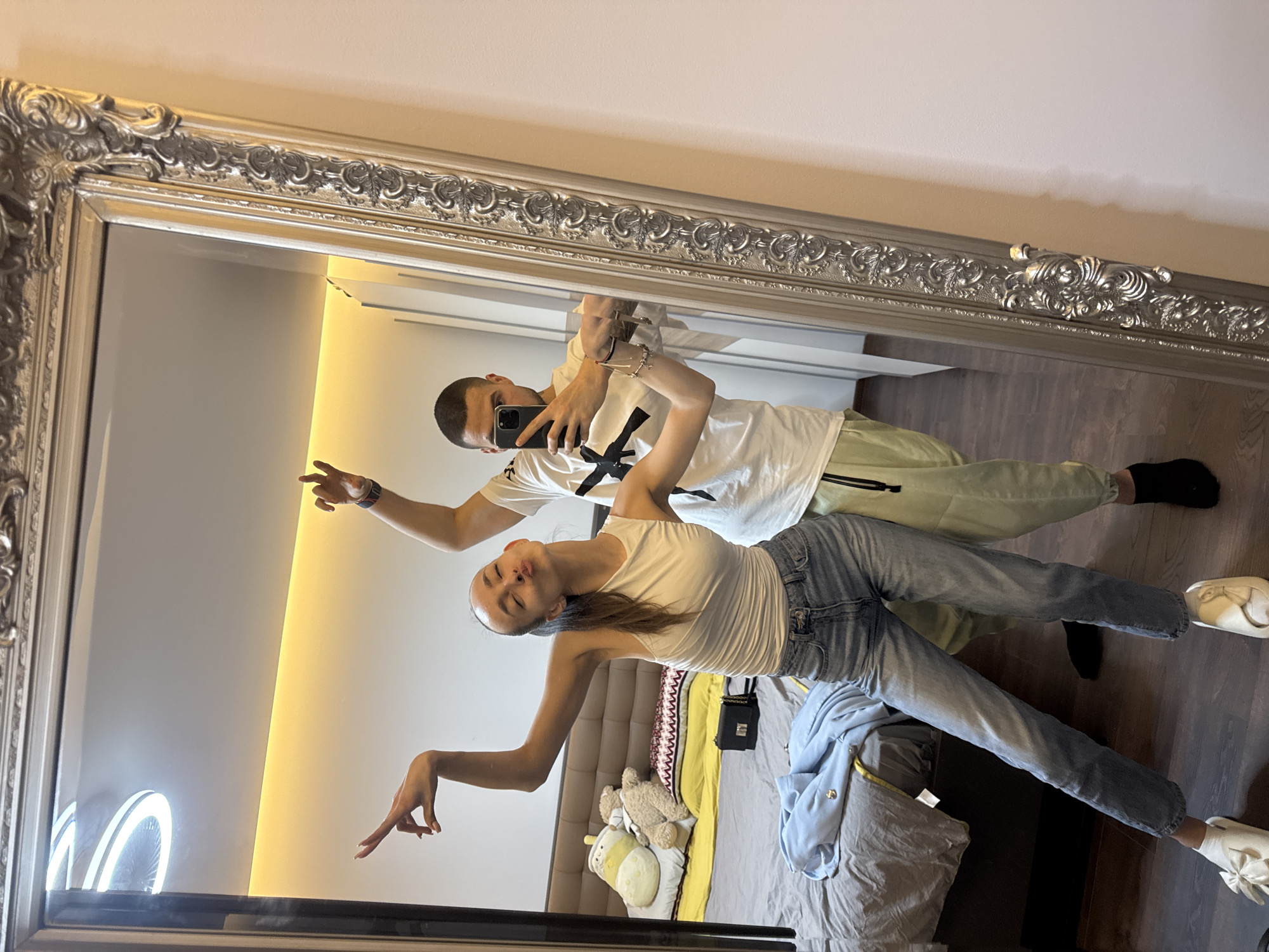 The image displays a mirror selfie of two individuals, a female and a male, within what appears to be a residential bedroom. The specific location in Sofia, Bulgaria, is not discernible from the visual information provided.
The female, positioned on the left in the reflection, wears a white sleeveless top, light blue denim jeans, and white slippers with bows. Her hair is pulled back, and she is striking a pose with both arms raised and bent, fingers extended, her eyes closed, and lips pursed. The male, positioned behind her, wears a white long-sleeved t-shirt with a black graphic on the chest and light green athletic pants. He is holding a dark-colored smartphone, consistent with an iPhone, with his right hand extended to capture the reflection. His left arm is also raised and bent. He has a short haircut, a dark wristband on his right wrist, and multiple bracelets on his left.
The room's interior, visible in the mirror's reflection, includes a large bed with a brown upholstered headboard, grey bedding, a yellow pillow, and white pillows. Resting on the bed are a yellow boba tea plushie, two brown teddy bears, a small black handbag with a silver chain, and a light blue garment. A linear LED strip light illuminates the wall above the bed, and a circular ceiling light fixture is visible in the upper left of the reflection. The floor is dark wood, and the walls are light-colored, with white paneled furniture or a door visible to the right of the bed. The mirror itself features an ornate, silver-colored frame with intricate carvings.