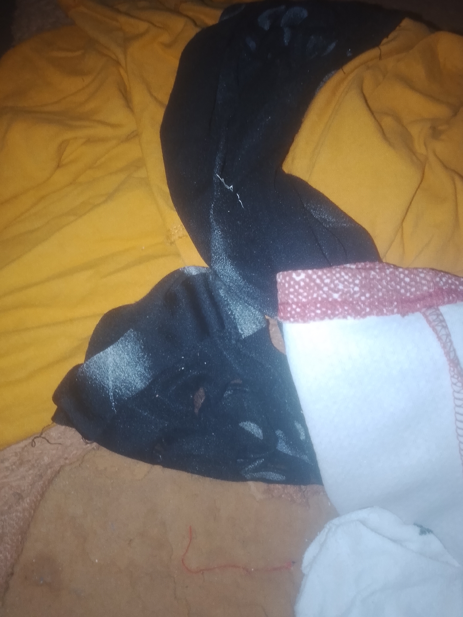 A close-up, overhead shot shows a pile of folded clothes. The dominant colors are yellow, black, and white, with hints of brown and red. The yellow fabric appears to be a t-shirt or similar garment, laid out flat. A black garment, possibly leggings or a skirt, is draped over the yellow fabric. A white garment, possibly underwear or a towel, with a red and white patterned trim, is placed on top of the black fabric. The background is a mix of brown and beige fabrics, possibly a carpet or blanket. There is a thin red thread visible on the brown fabric. The lighting is dim, and the image has a slightly grainy texture. The scene appears to be indoors, possibly in a bedroom or laundry area, within Jalingo, Nigeria. There are no people visible in the image.