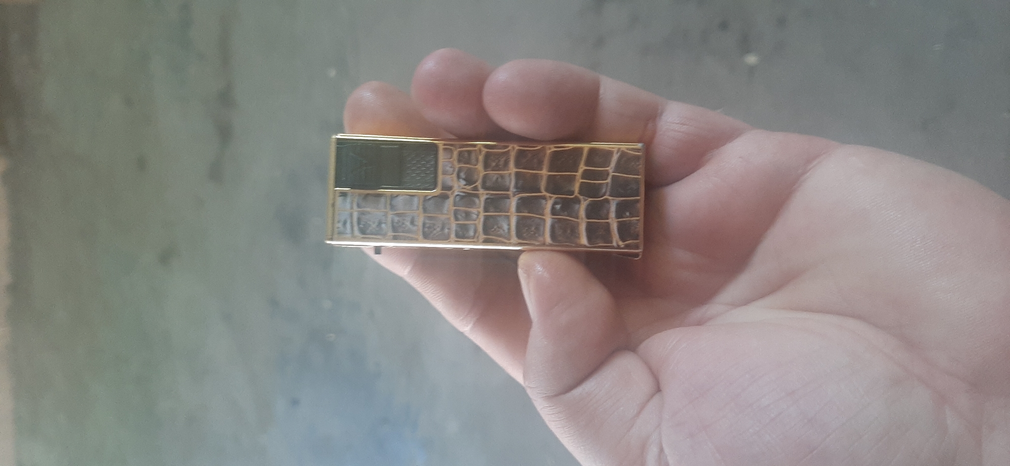 A close-up, angled overhead shot shows a person's hand holding a rectangular gold-colored object with a crocodile-skin pattern. The object has a green rectangle on its left side. The background is a blurred, gray, textured surface, possibly concrete or asphalt. The lighting is diffused, suggesting it might be daytime, but no specific time of day can be determined. The image is taken in Yessentukskaya, Russia.