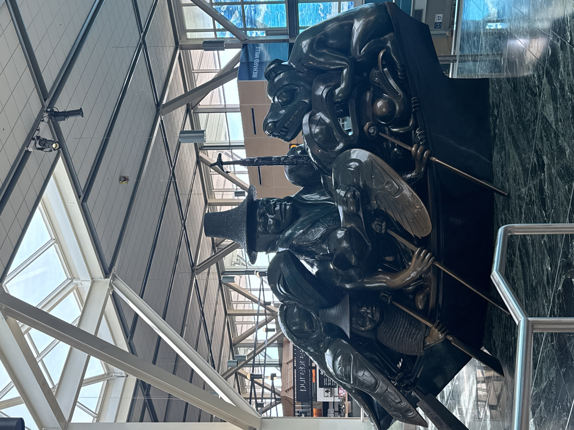 This image depicts a bronze sculpture located inside the Vancouver International Airport (YVR) in Richmond, Canada. The sculpture features a bear-like figure on top, and a man in a top hat is located beneath it. The sculpture has multiple figures and details. The surrounding environment includes the airport's architectural elements, such as a patterned ceiling with metal support beams, glass windows, and marble-like wall panels. In the background, there is signage for airport amenities. A railing is visible on the right side of the image.