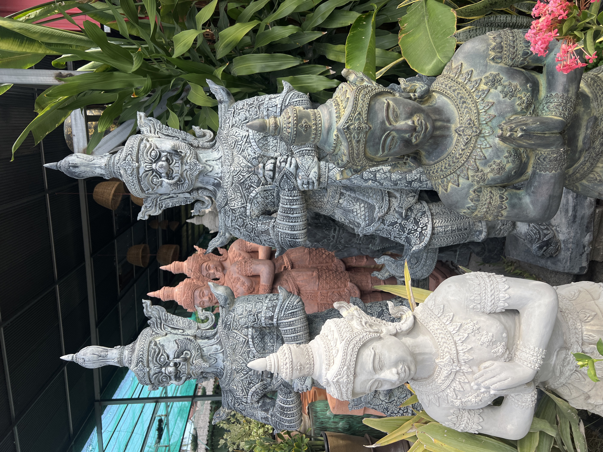 The image depicts an outdoor display of several large stone statues in what appears to be a garden or marketplace in the Ko Pha-ngan District of Thailand. In the foreground, a white stone statue of a figure with hands pressed together in a prayer-like gesture faces the viewer. To its right, a weathered bronze-colored statue of a similar figure with folded hands is visible, adorned with intricate jewelry. Behind these, several imposing gray statues of figures with fierce expressions and elaborate headdresses stand tall, some holding objects. In the middle ground, two reddish-brown statues of figures with similar headdresses and hands pressed together are partially obscured. The background shows a covered structure, possibly a roof or awning, with a turquoise tarp to the left and lush green foliage to the right, including large, broad leaves. Woven lampshades hang from the ceiling of the structure. The overall scene is of a vendor displaying religious or decorative statuary.