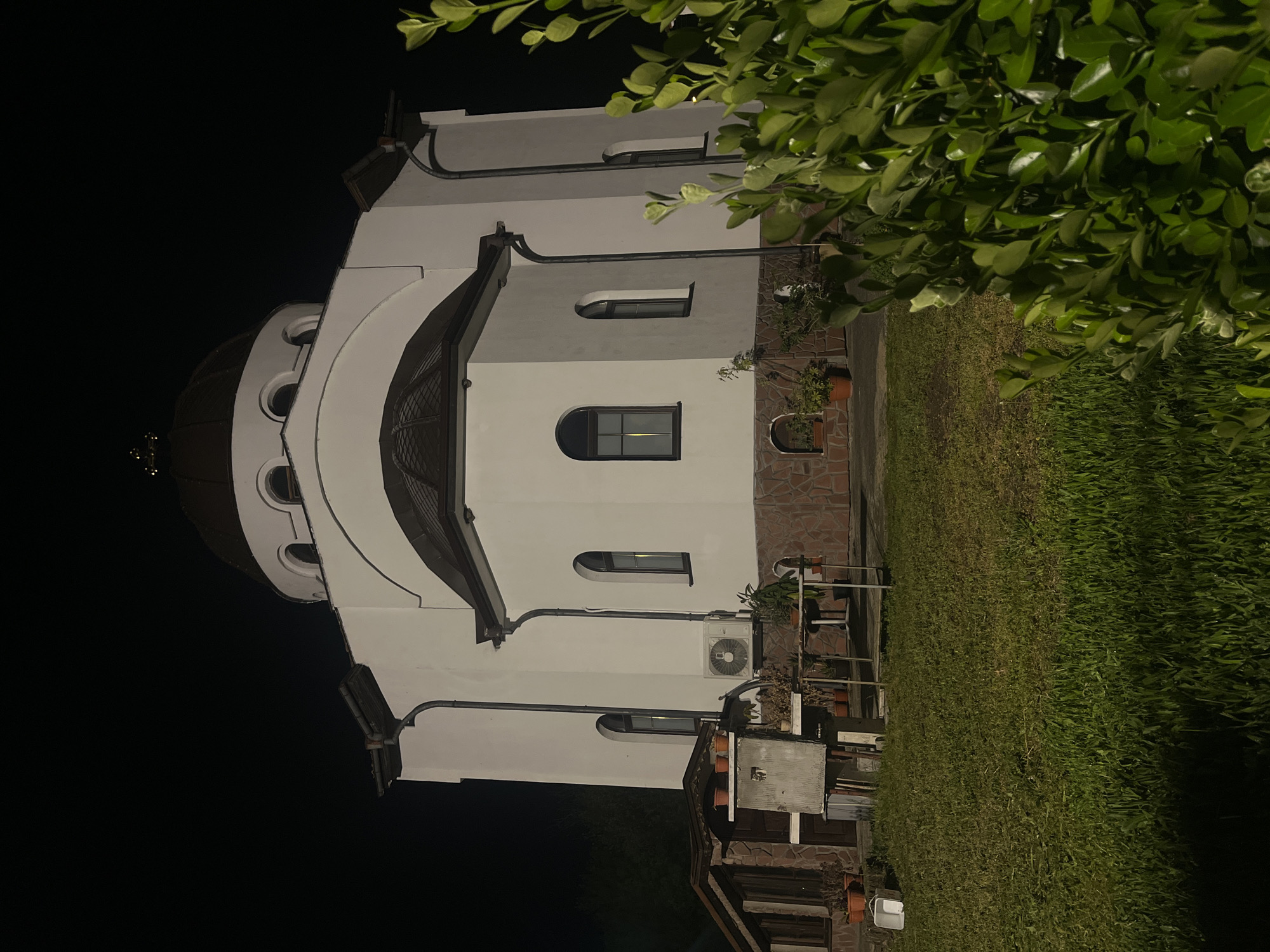The image depicts a white building with a dome at night. The building has arched windows and a stone base. There is a patch of green grass adjacent to the building, and some foliage visible on the right side of the image. The left side of the image is mostly dark. Based on the architecture and information available online, this building appears to be the Church of St. Nicholas of Myra in Sofia, Bulgaria.