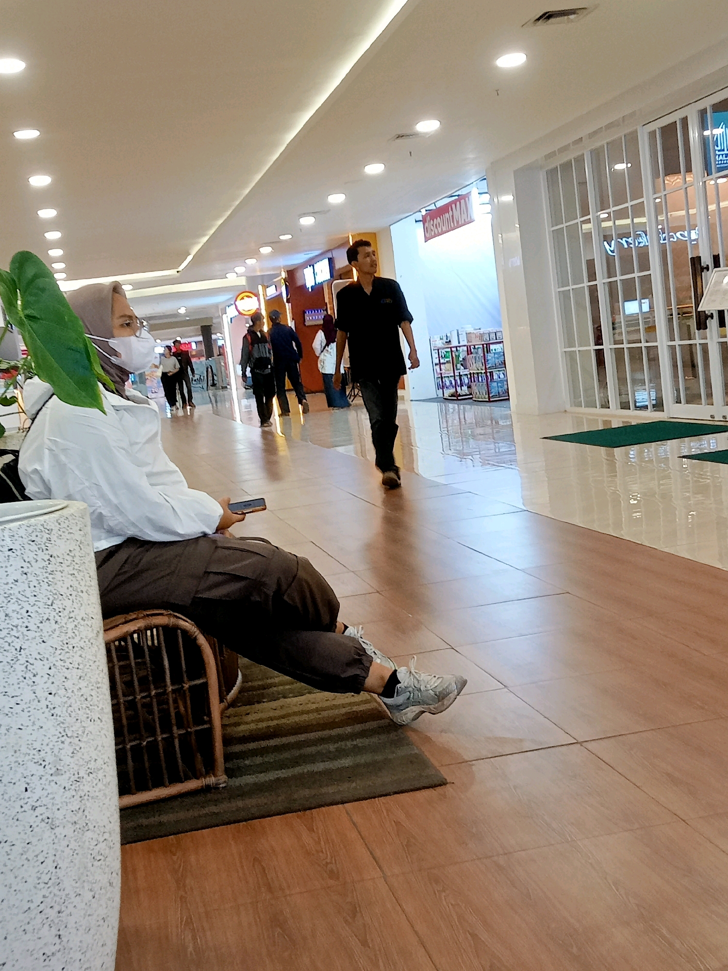 The image depicts an indoor scene, likely a shopping mall or commercial complex, in Cirebon, Indonesia. In the foreground, a person wearing a hijab, mask, white jacket, and brown pants is seated on a rattan chair, holding a smartphone. To the right, a man in a black shirt and dark pants walks away from the camera. The environment features a polished floor with a wood-grain tile pattern, a white ceiling with recessed lighting, and a large glass storefront with a white grid pattern. Signs visible in the background include "discountMAX". Other individuals are seen walking in the distance. The overall atmosphere suggests a public, commercial space.