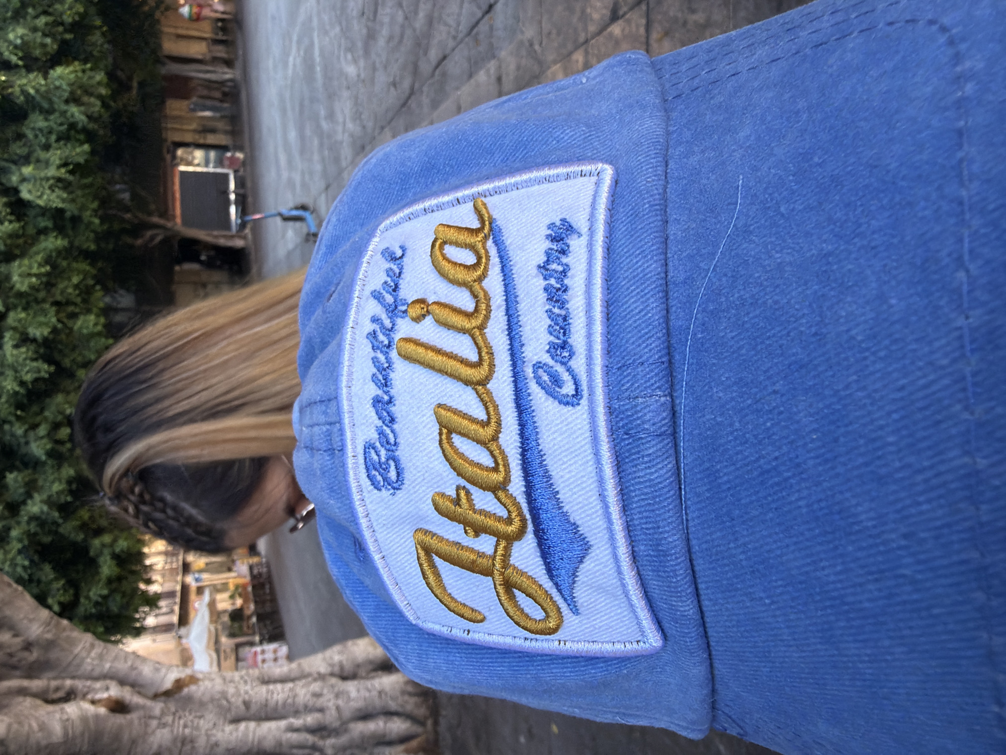 The image is a close-up of a light blue baseball cap worn by an unseen person, with the back of their head and blonde hair visible. The cap features an embroidered patch with the words "Beautiful Italia Country" in gold and blue lettering. The background is blurred, showing trees, buildings, and a paved area, suggesting an outdoor urban setting. The overall impression is that of a tourist souvenir cap.