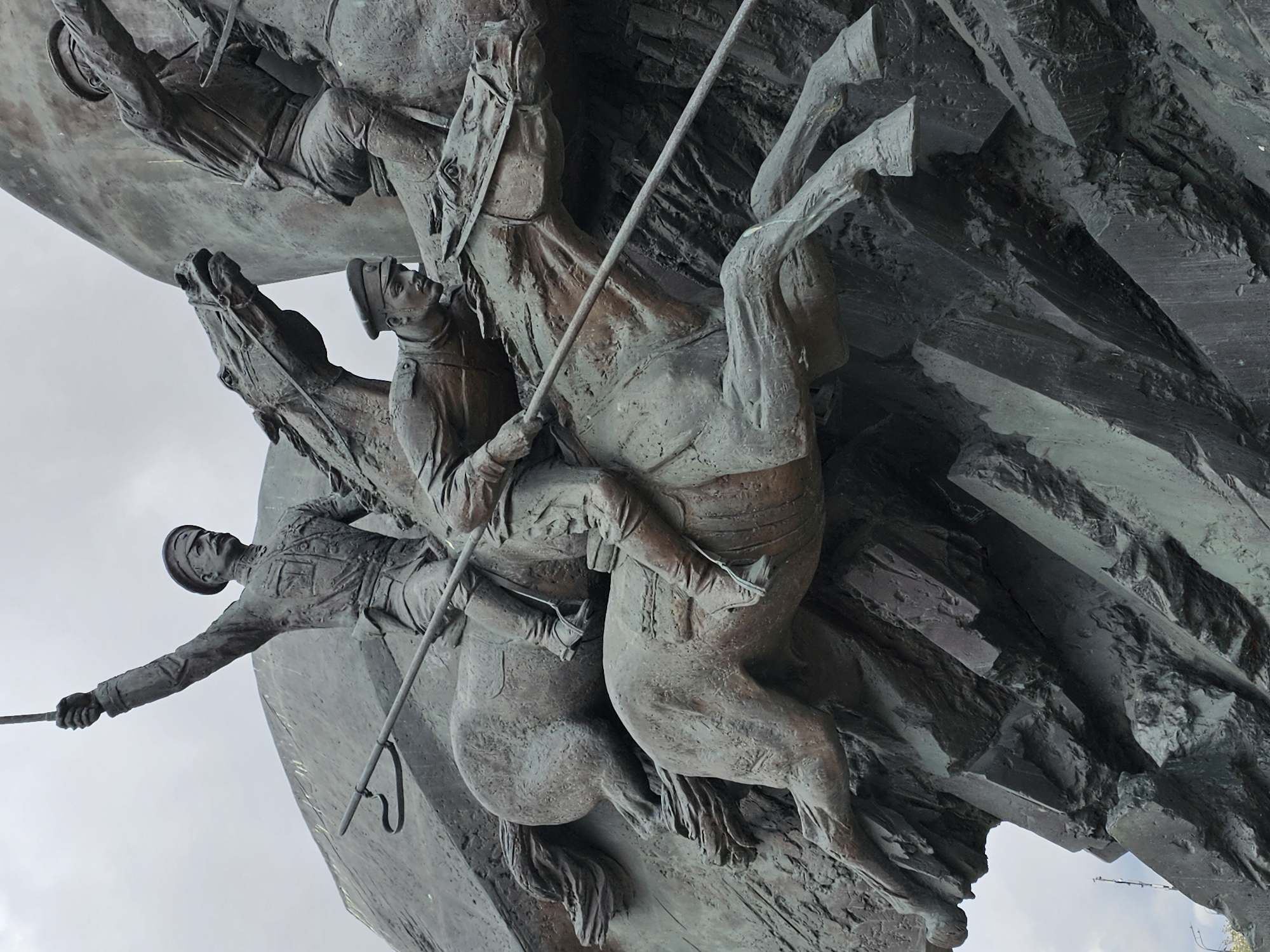 The image captures a bronze sculpture of soldiers on horseback, a detail from the Monument to the Heroes of World War I, located on Poklonnaya Hill in Moscow, Russia. The monument depicts several mounted soldiers, clad in period uniforms, equipped with swords and spears. The sculpture is mounted on a stone base, which seems to rise up into a jagged, rock-like formation. The sky is visible in the background.