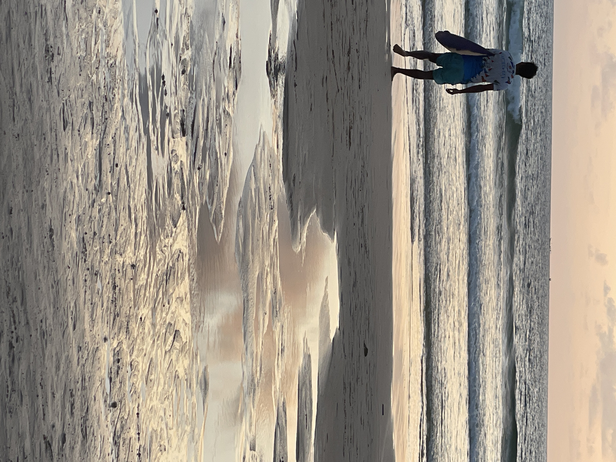 A lone individual, a male with dark hair, walks barefoot along a sandy beach in Rawai, Thailand. He is dressed in a patterned short-sleeved shirt and blue shorts, carrying a surfboard under his left arm. The man is facing away from the camera, walking towards the ocean. Gentle waves are breaking on the shore, and the water extends to the horizon where a pale, cloud-streaked sky meets the sea. The sand in the foreground shows the texture of wet sand with puddles reflecting the sky's light. The overall scene depicts a tranquil beach setting during what appears to be either sunrise or sunset.