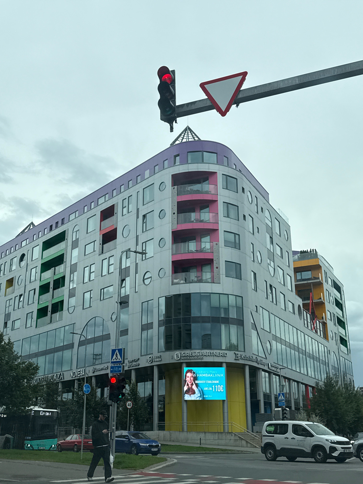 This is a photograph taken from street level, looking up at a modern, multi-story building with a curved facade. The building is predominantly clad in grey metal panels, with colorful accents of purple at the top, pink balconies, and green sections on the facade. The windows are a mix of rectangular and circular shapes.
In the foreground, a traffic light displays a red signal. A street sign with a blue pedestrian crossing symbol and a bicycle symbol is visible below it. A person wearing a black hooded sweatshirt and black pants is walking across a crosswalk. Several vehicles are present: a turquoise bus, a red car, a dark blue car, and a white van.
Above the building, a traffic sign displaying a red-bordered white triangle, commonly known as a "yield" or "give way" sign, is suspended from a metal bar. The sky is overcast and appears to be a light grey.
The building appears to be part of a commercial and residential complex. Signs on the building include "ORION PLAZA", "NOVEBA", "LaserMed", and "EESTI FIRMA". A large digital advertisement screen displays an image of a woman with the text "Kadrioru Plaza HAMBAKLIINIK HAMBAKIVI EEMALDAMINE AIRFLOW 110€". This indicates the location is in Tallinn, Estonia, likely in the Kadriorg district, given the advertisement.