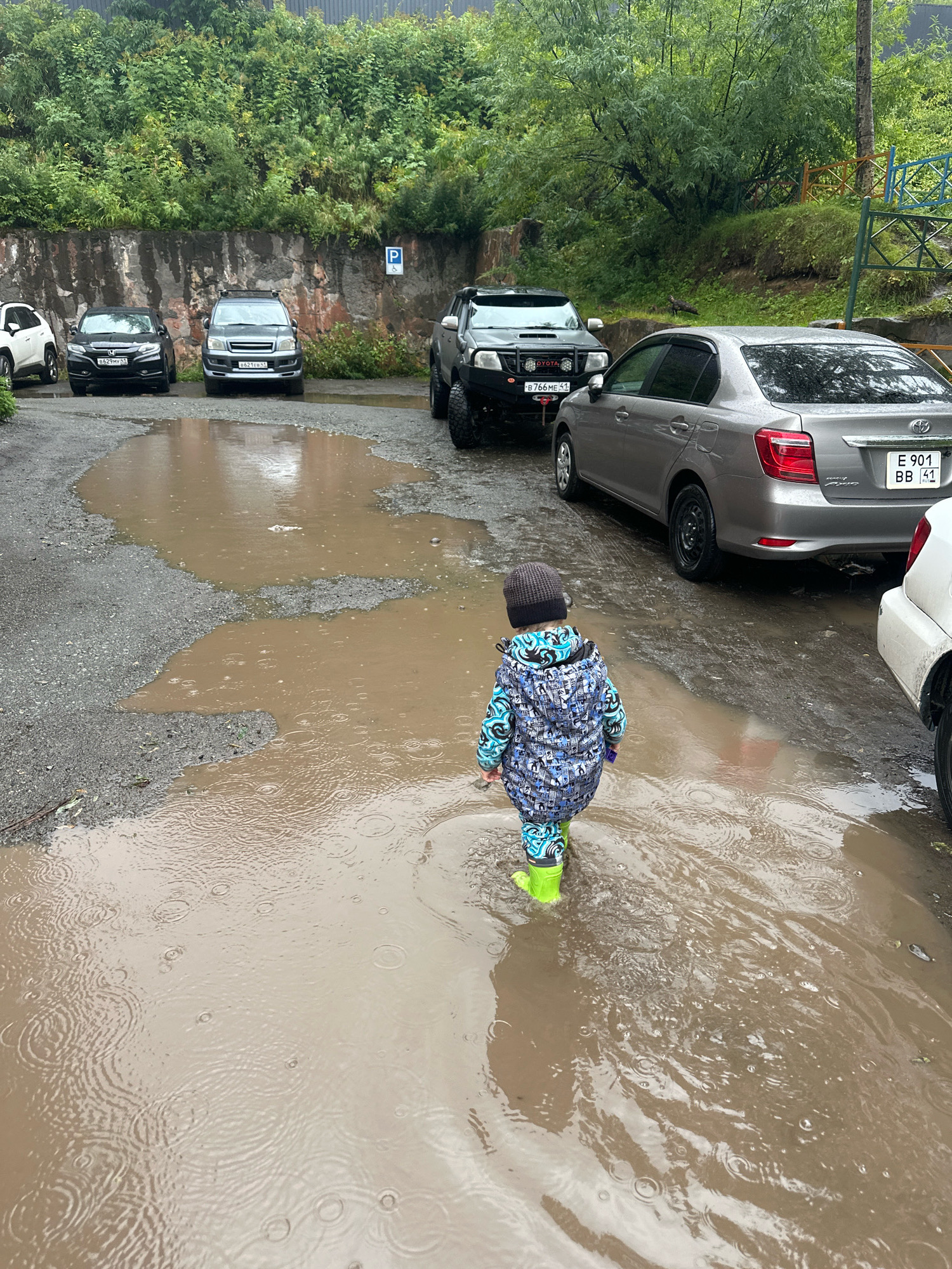 A toddler wearing a grey knitted hat, a blue and black patterned jacket, and bright green rubber boots walks away from the camera through a large, muddy puddle on a gravelly, wet surface. The puddle shows ripples from rainfall. In the background, several vehicles are parked, including a silver sedan with a license plate reading "E 901 BB 41", a black Honda, a dark grey SUV with a bull bar, and a white vehicle partially visible on the right. Behind the cars, a rough stone wall is visible, covered in green foliage and trees. A blue and white parking sign with a "P" is affixed to the wall. The environment appears to be outdoors in a city, possibly a parking area, with evidence of recent rain.