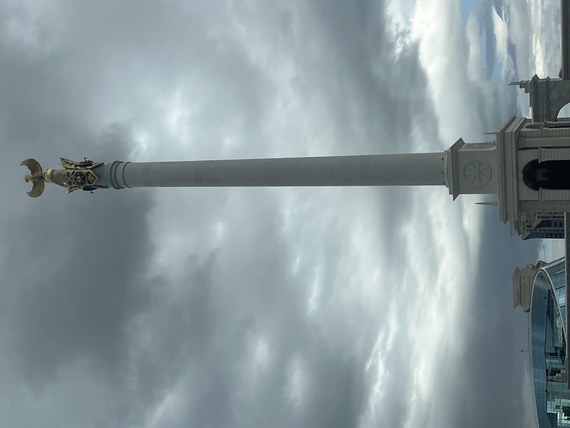 The image shows the Bayterek Tower in Astana, Kazakhstan, under a dramatic, cloudy sky. The tower is a prominent landmark, characterized by its tall, slender white column topped with a golden, eagle-like sculpture. The base of the tower is ornate, featuring architectural details and decorative elements. In the foreground, the sleek, modern architecture of surrounding buildings is visible, including a structure with a curved, glass facade. The overall scene is one of urban grandeur set against a moody, atmospheric backdrop.