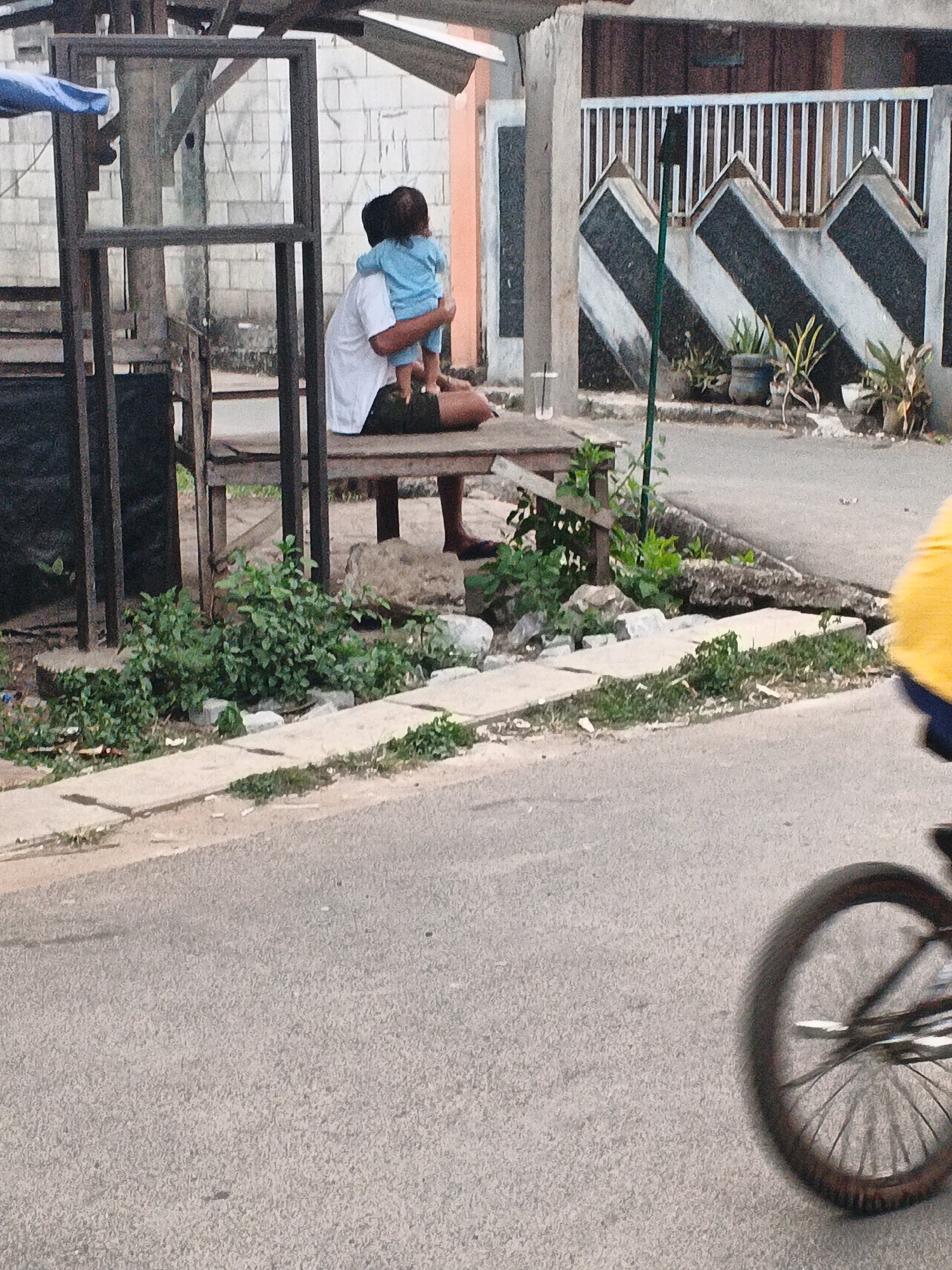 In Kadu, Indonesia, a man in a white shirt and dark shorts sits on a wooden bench, holding a child in a blue shirt on his lap. The child faces away from the viewer. Behind them, a rough brick wall is visible, partially obscured by a wooden structure with vertical supports. To the right of the man and child, a street is visible with a grey asphalt surface. In the foreground, on the right side of the image, a portion of a bicycle wheel and a person in a yellow shirt are in motion, suggesting movement on the street. Green plants and some debris are scattered on the ground near the bench. A decorative fence with a zig-zag pattern is visible in the background.