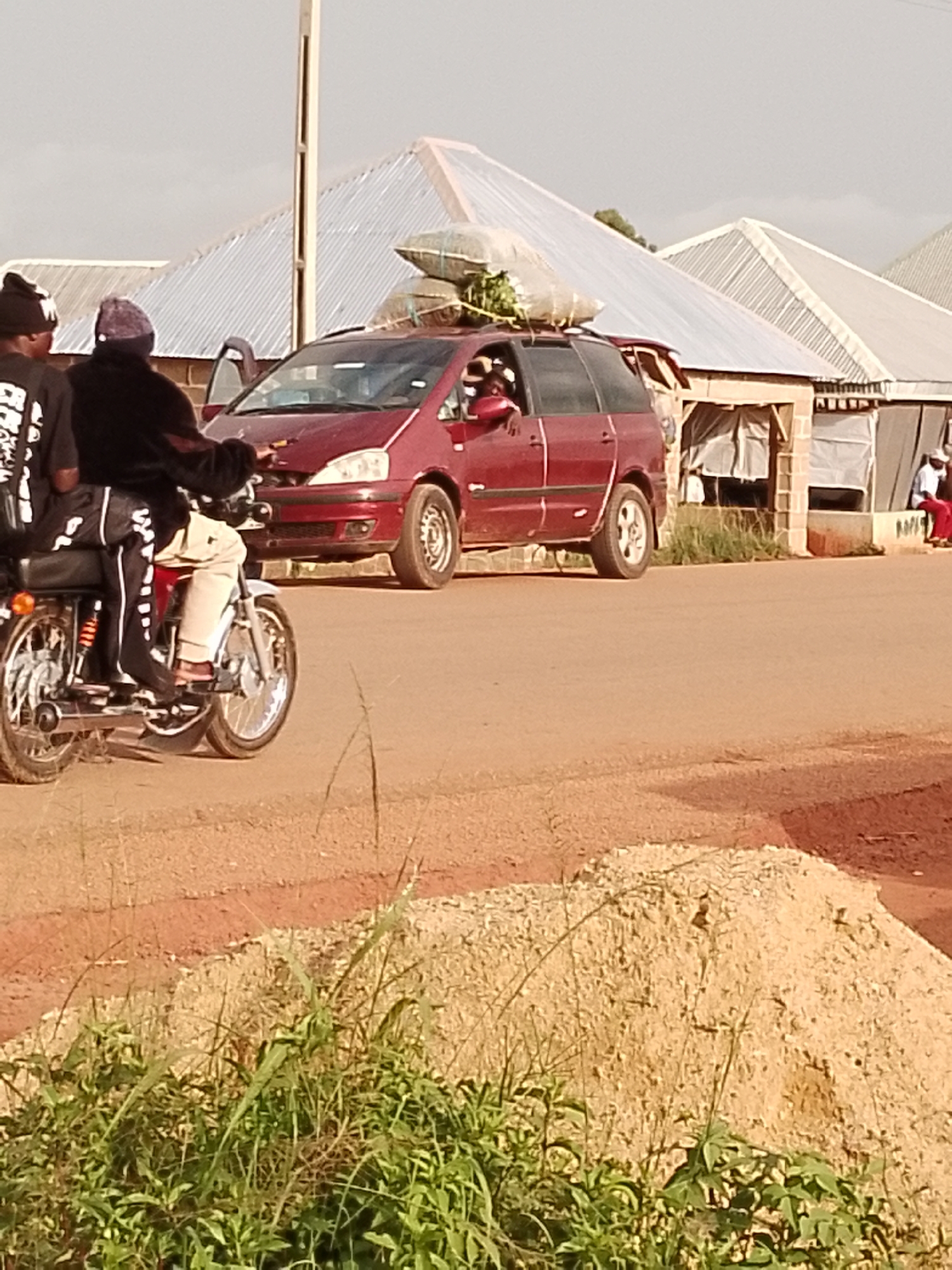 In this image, two individuals are seen on a motorcycle, with one driving and the other as a passenger. The driver is wearing a black hooded jacket and a beanie, while the passenger is wearing a black t-shirt with white lettering and a beanie. They are on a dirt road in what appears to be a rural or semi-urban setting.
Behind the motorcycle is a red minivan with its front passenger door open. The minivan is loaded with goods on its roof, including what looks like bundled plants or produce, secured with ropes. A person can be seen leaning out of the driver's side window of the minivan.
In the background, there are several buildings with corrugated metal roofs, suggesting a developed area. Some of these buildings have brick walls and open fronts covered with white sheeting, possibly indicating shops or market stalls. Another person in red pants and a white shirt is visible sitting in front of one of these structures. The sky is overcast, suggesting it might be a cloudy day. The overall scene depicts a busy, practical moment, likely involving transportation of goods and people in Agwala, Nigeria. There is no visible text in the image.