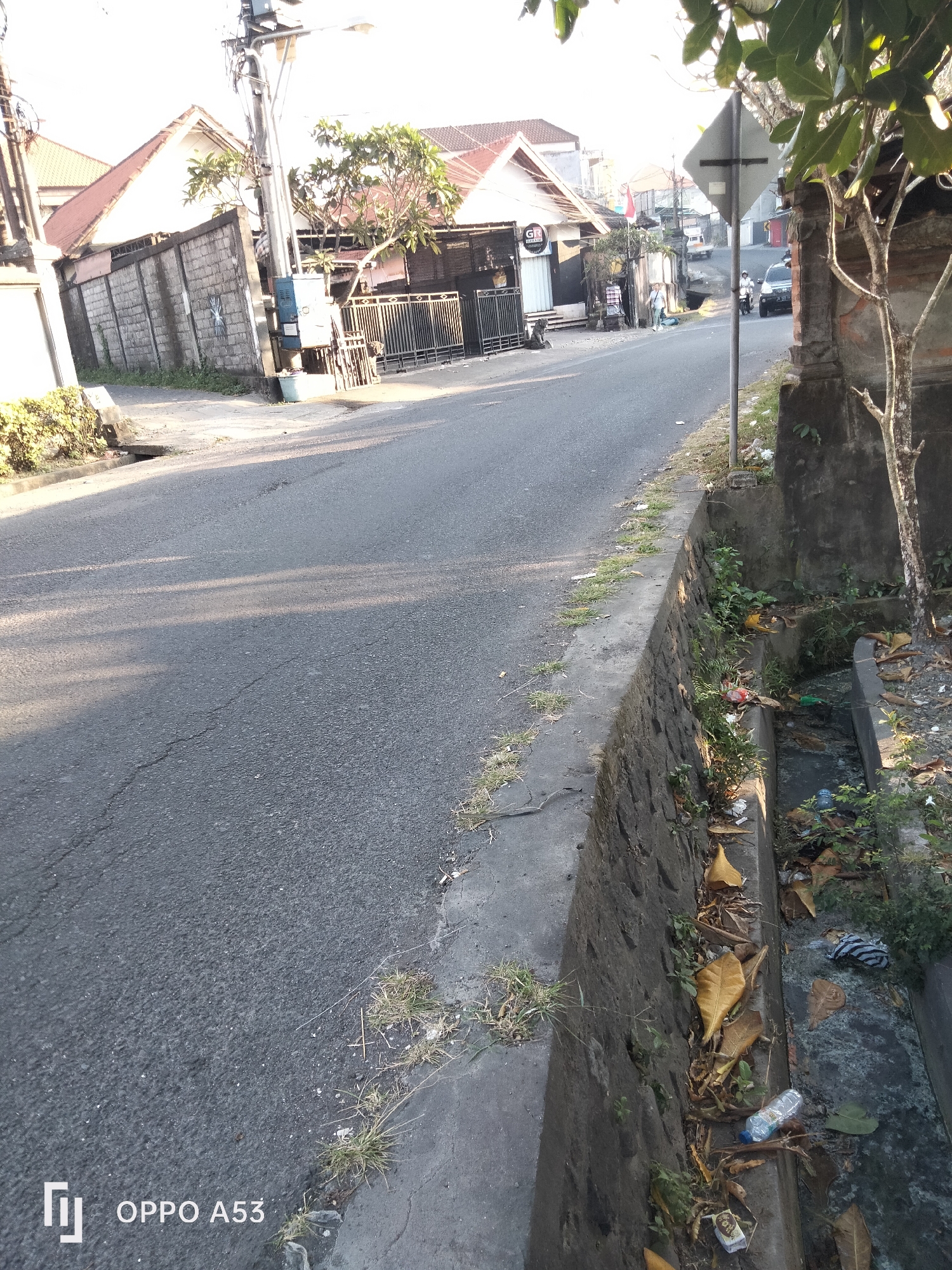 The image depicts a street scene in Denpasar, Indonesia. A paved road occupies the left and center of the frame, with a textured asphalt surface. To the right of the road is a concrete drainage ditch filled with debris, including fallen leaves, plastic bottles, and other litter. A stone wall borders the ditch, and vegetation grows along its edge and within the ditch itself. On the left side of the road, there is a low concrete wall and a patch of grass. Beyond the road and wall on the left are buildings with red tile roofs and a concrete fence. A utility pole with wires and a street lamp is visible on the left side of the road. In the background, more buildings and a street with a parked car and a motorcycle can be seen. The lighting suggests it is daytime, with shadows cast across the road. The "OPPO A53" watermark is present in the bottom left corner.