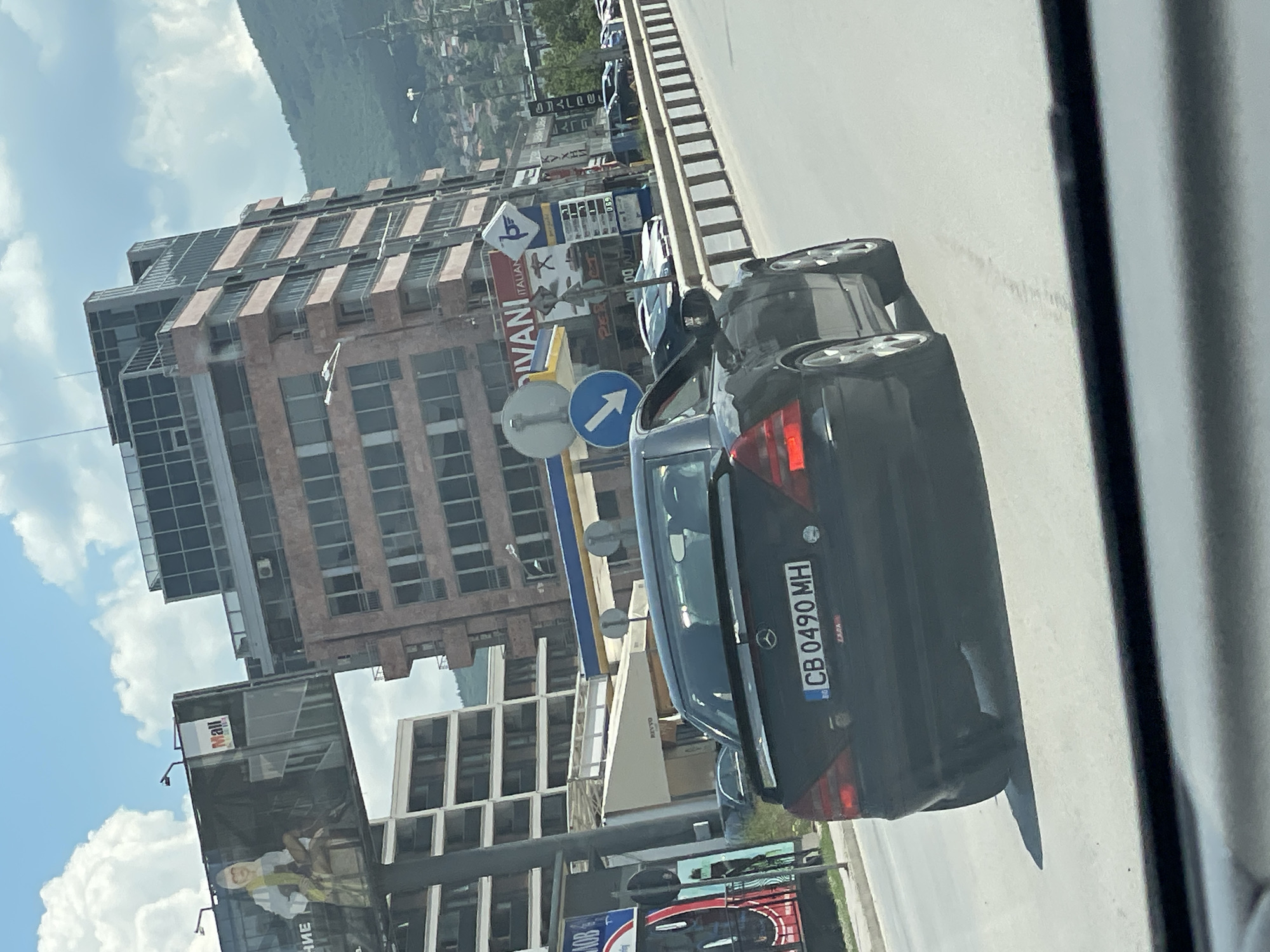 The image shows a black Mercedes-Benz SLK convertible with license plate CB 0490 MH, driving on a road in Sofia, Bulgaria. The car is in the foreground, and its rear is visible. In the background, there are several buildings, including a modern office building with glass facades and a billboard advertising "Mall". There are also some smaller buildings and a gas station visible. The sky is cloudy, and a mountain range is visible in the distance. The scene appears to be captured during the daytime.
