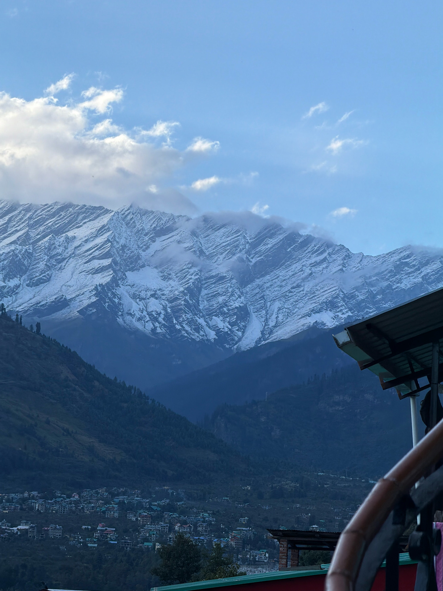 A wide-angle shot captures a panoramic view of snow-capped mountains in Vashisht, India. The mountains are majestic, with their peaks dusted in white snow, contrasting against the clear blue sky dotted with fluffy white clouds. A village is nestled at the foot of the mountains, with buildings and houses clustered together. The foreground features a partial view of a rooftop or balcony, suggesting the photo was taken from a higher vantage point. The overall atmosphere is serene and breathtaking, evoking the natural beauty of the Himalayas.