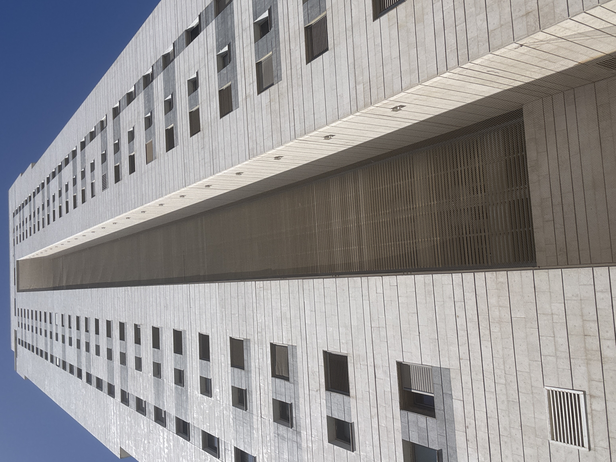 A low-angle shot of a tall, multi-story building with a clear blue sky as the backdrop. The building, likely located in Netanya, Israel, features a uniform pattern of windows and a light-colored, possibly concrete or stone, facade. A large horizontal section spans a significant portion of the building's width. The image is tilted, giving the viewer the impression of looking upwards at the structure.