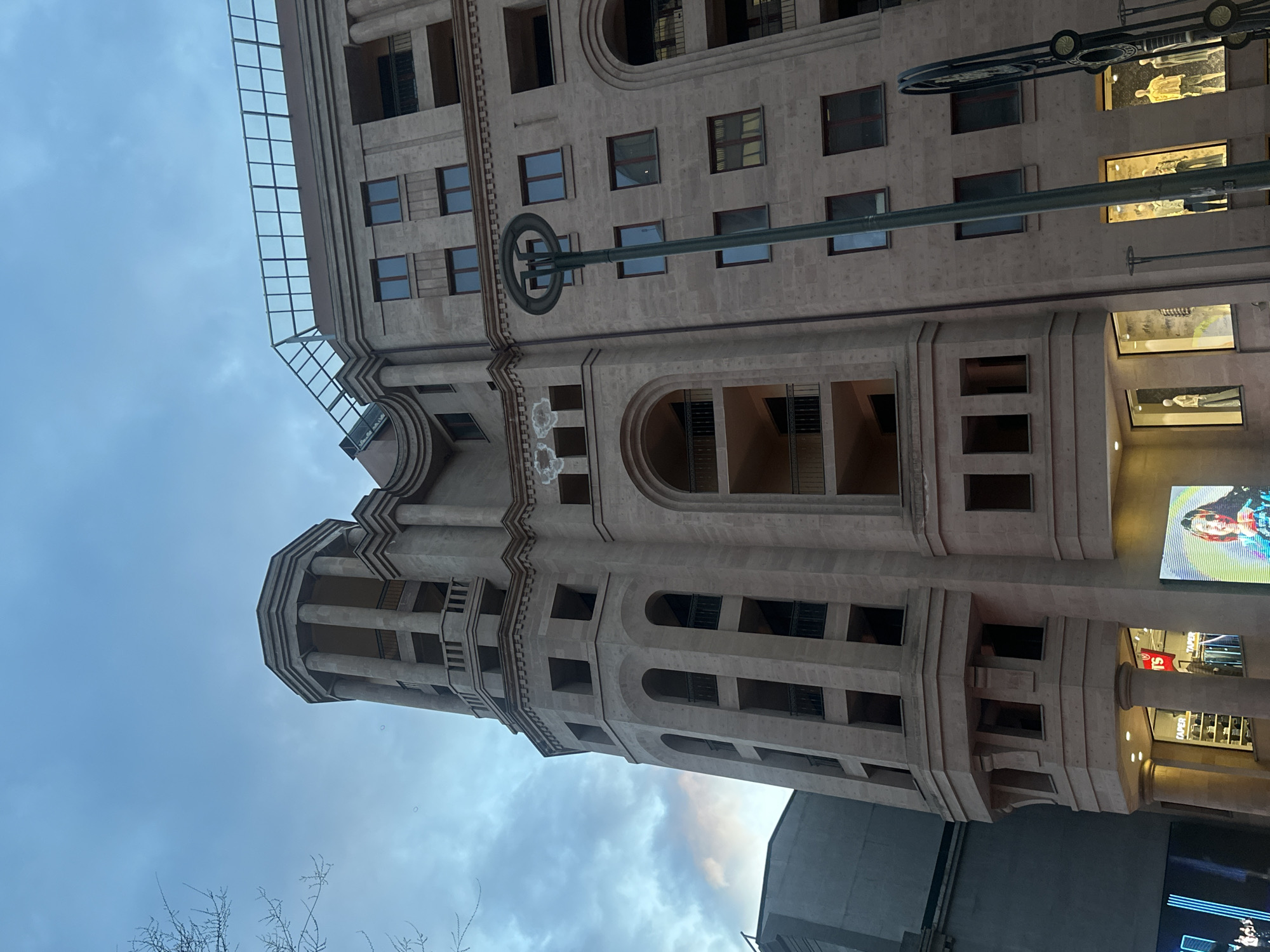 The image depicts a tall, multi-story building with a light-colored stone facade in Yerevan, Armenia. The architectural style features arched windows, columns, and balconies. The building appears to be commercial, with display windows and possibly shops at the ground level. A street lamp extends horizontally in front of the building, with circular decorative elements. The sky is overcast, suggesting either early morning or late afternoon. The overall impression is of a modern building with classical architectural influences.