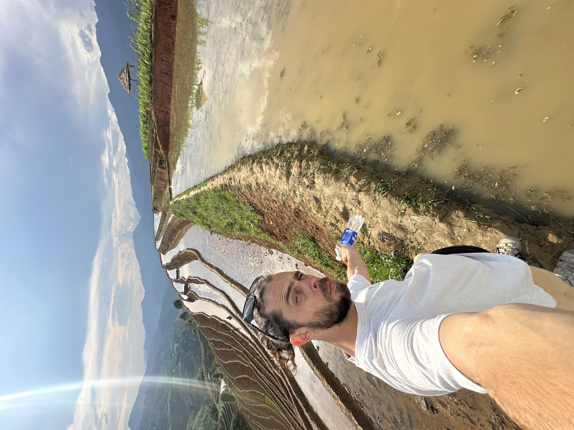 The image captures a man taking a selfie or being photographed amidst terraced rice fields in Lao Chai, Vietnam. The man, with a beard, dark curly hair, and sunglasses pushed onto his head, wears a white t-shirt. He is holding a clear plastic water bottle with a blue label that reads "Aqua" in his right hand, extended towards the camera. He looks slightly upwards and to his right, with a calm expression.
The setting is a vibrant agricultural landscape characterized by numerous terraced rice paddies, which descend down a mountainous slope. Many of the paddies are filled with water, reflecting the bright blue sky and scattered white clouds above. The water in some of the lower terraces appears murky brown with visible debris and small plants, while higher terraces show clearer reflections.
In the background, majestic mountains rise under a clear sky, indicating good weather. A small, rustic hut with a thatched roof is visible on a distant ridge, adding a touch of local architecture to the natural scenery. The overall impression is one of a vast, tranquil, and picturesque rural environment.
The time of day appears to be bright daytime, likely late morning or early afternoon, given the strong sunlight and clear skies, with a subtle lens flare suggesting direct sun exposure. The scene conveys a sense of peaceful exploration or appreciation of the scenic surroundings.