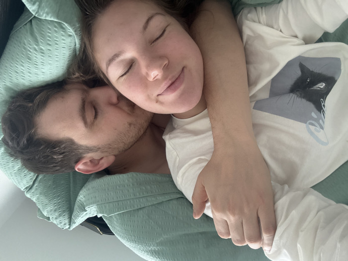 The scene depicts two individuals, one female and one male, in close physical proximity, appearing to be asleep or resting in a bed within an indoor setting in Vozdovac Urban Municipality, Serbia.
The female is positioned on her back, her head resting on a white pillow, tilted slightly upwards and to the left. Her eyes are closed. She has medium-brown hair. She is wearing a white long-sleeved top featuring a graphic of a black cat's head with white eyes, overlaid with a light blue geometric design. Her left arm is extended and positioned across the male's back.
The male lies on his right side, facing left, with his head resting against the female's left shoulder and upper chest. His eyes are closed. He has short, dark brown hair and visible facial stubble. He appears to be shirtless or wearing a light-colored undershirt, largely concealed by the bedding.
Both individuals are covered by a mint-green, quilted duvet or blanket. A dark, rectangular object, possibly an electronic device or small bag, is partially visible behind the male's head, resting on the duvet. The lighting suggests ambient light from an unseen source.