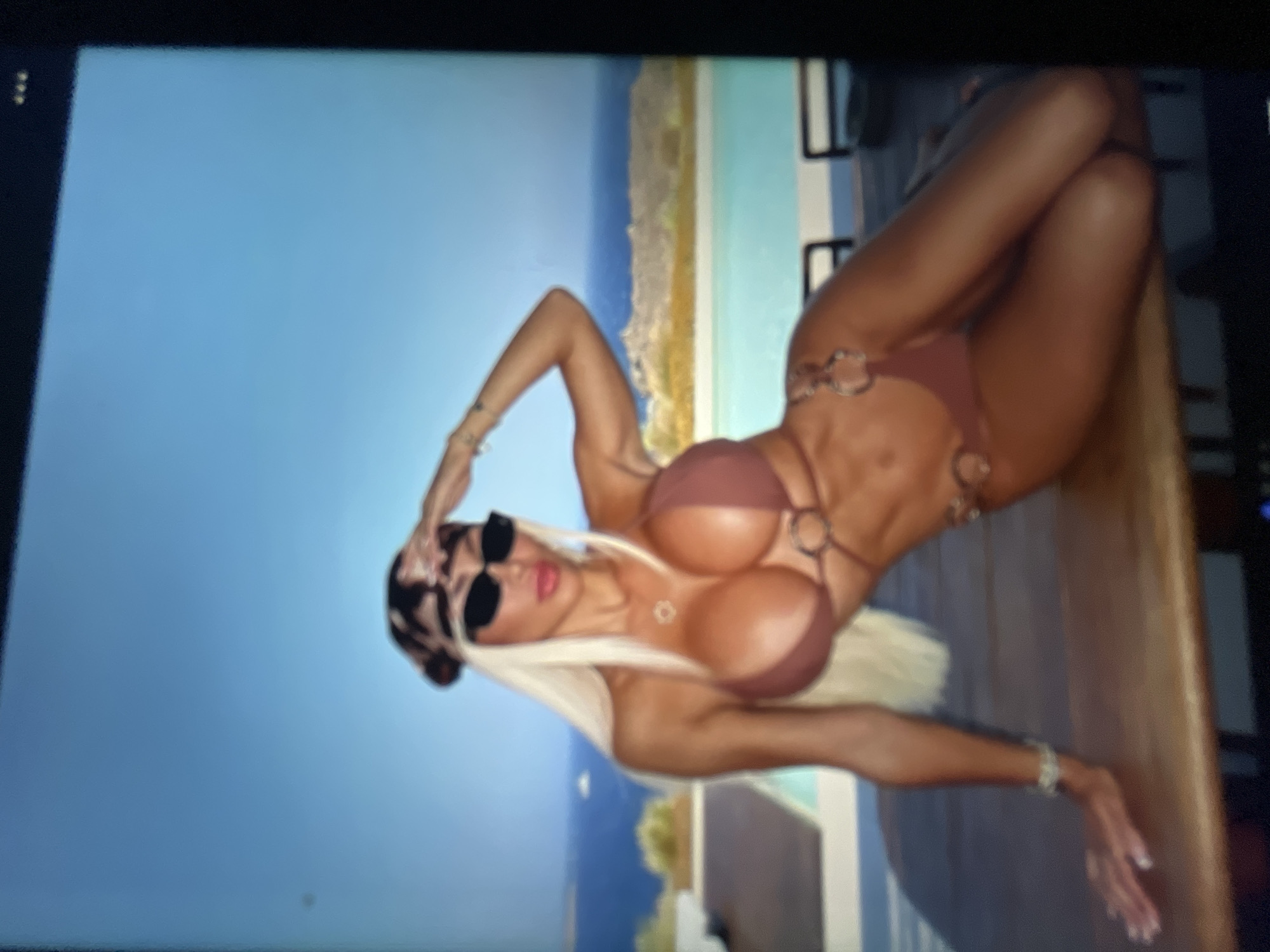 The image displays a woman in a brown bikini posing on a wooden surface. She has long blonde hair, wears sunglasses and a bandana. Behind her, there is a body of water with islands and a clear blue sky. The scene appears to be outdoors, possibly a vacation spot, with a poolside setting. There is no indication that this location is in Sofia, Bulgaria.
