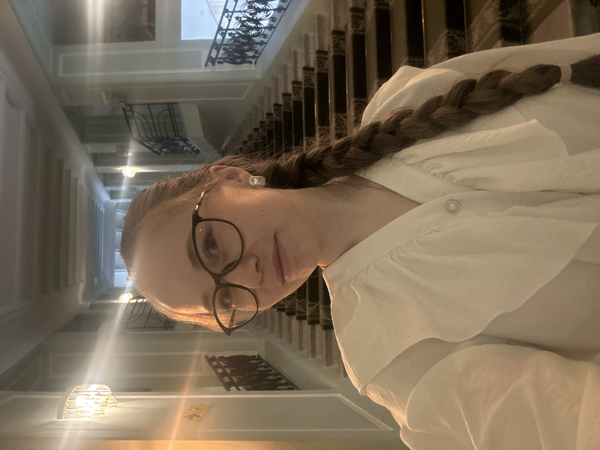The image shows a person wearing glasses and a white shirt with a braid, situated in a building that appears to be a public or historical venue. The setting seems to be Saint Petersburg, Russia, evidenced by the classical architectural elements visible in the background such as detailed railings and ornate interior design. The location appears to be an upper level with views into lower floors, suggesting it could be a stairwell.