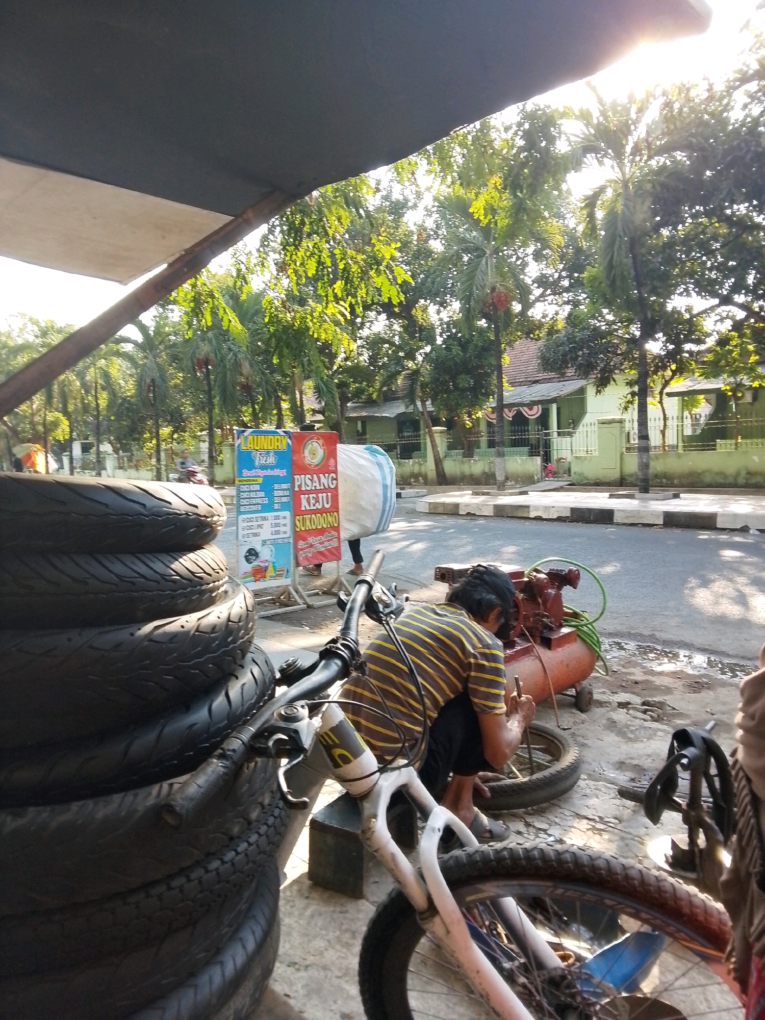 In Surabaya, Indonesia, under a sunny sky, a person wearing a striped shirt and dark pants is working on a tire on the side of a road. They are kneeling next to an orange air compressor with green hoses. In the foreground, several black tires are stacked vertically. A bicycle with white handlebars and frame is visible, partially obscured by the tires. Behind the worker, a sign displays "LAUNDRY FRESH" and "PISANG KEJU SUKODONO". The background shows houses with green walls and trees.