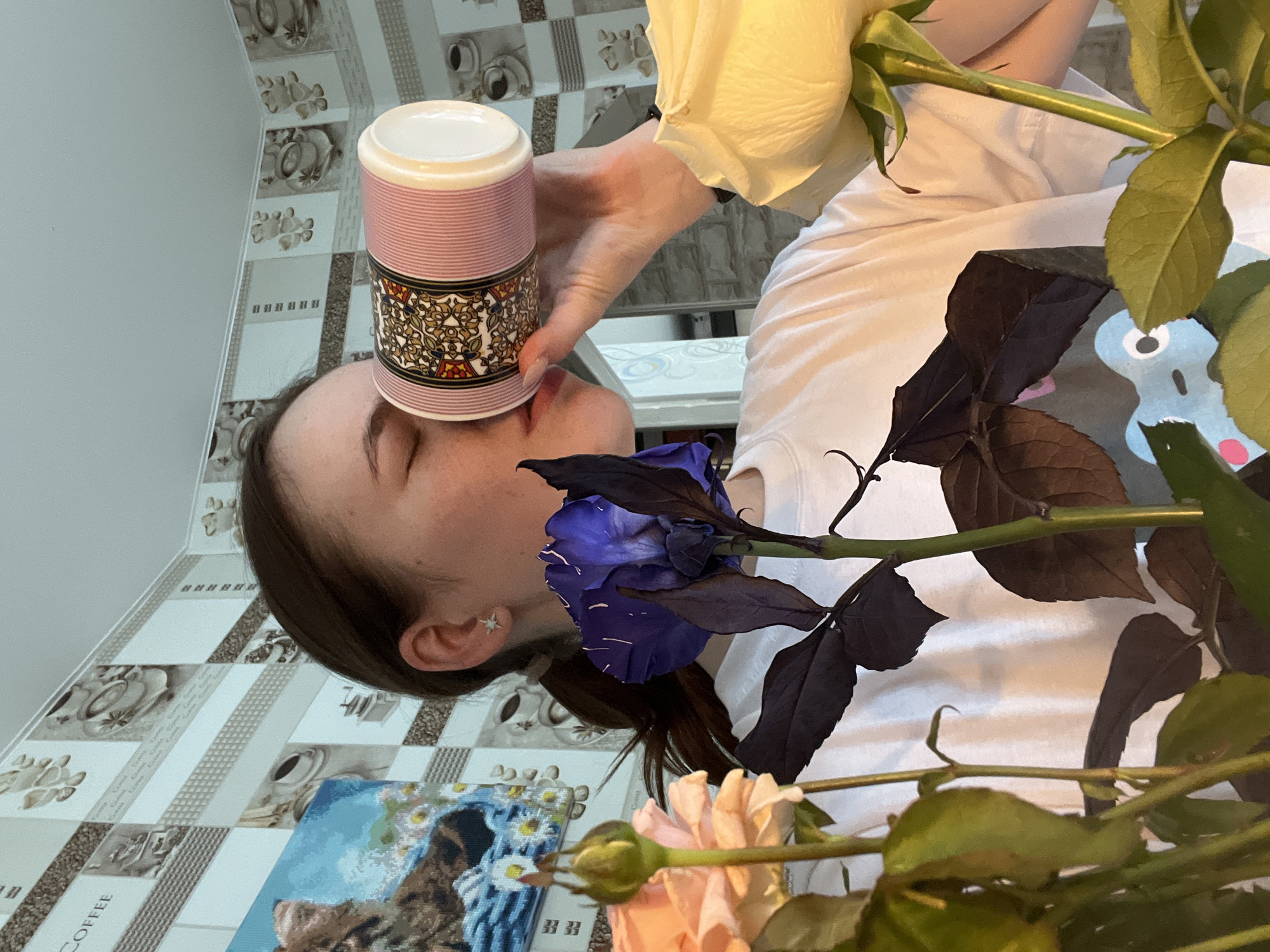 The image shows a person with their eyes closed, holding a pink and patterned cup against their forehead. A bouquet of roses in various colors (white, blue, peach) is positioned in the foreground. The person is wearing a white shirt. The background features a tablecloth with a coffee-themed pattern. A partially visible painting is present on the left side. The location is Chaikovsky, Russia.