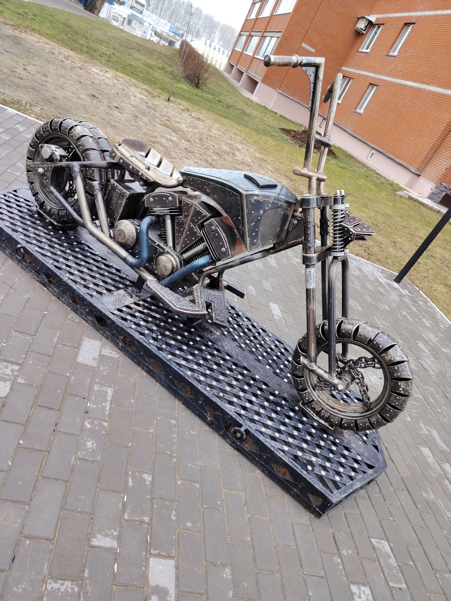 The image shows a metal sculpture of a motorcycle displayed outdoors in Chaikovsky, Russia. The motorcycle is composed of metal components such as tubes, plates, and chains, mimicking the appearance of a chopper-style motorcycle. It features studded tires, a riveted body, and high-rise handlebars. The sculpture is mounted on a black metal grating platform, which is set on a paved brick surface. In the background, there is a grassy area, a multi-story brick building with visible windows, and trees. The weather appears overcast, affecting the natural light in the scene. No people are visible in the image.