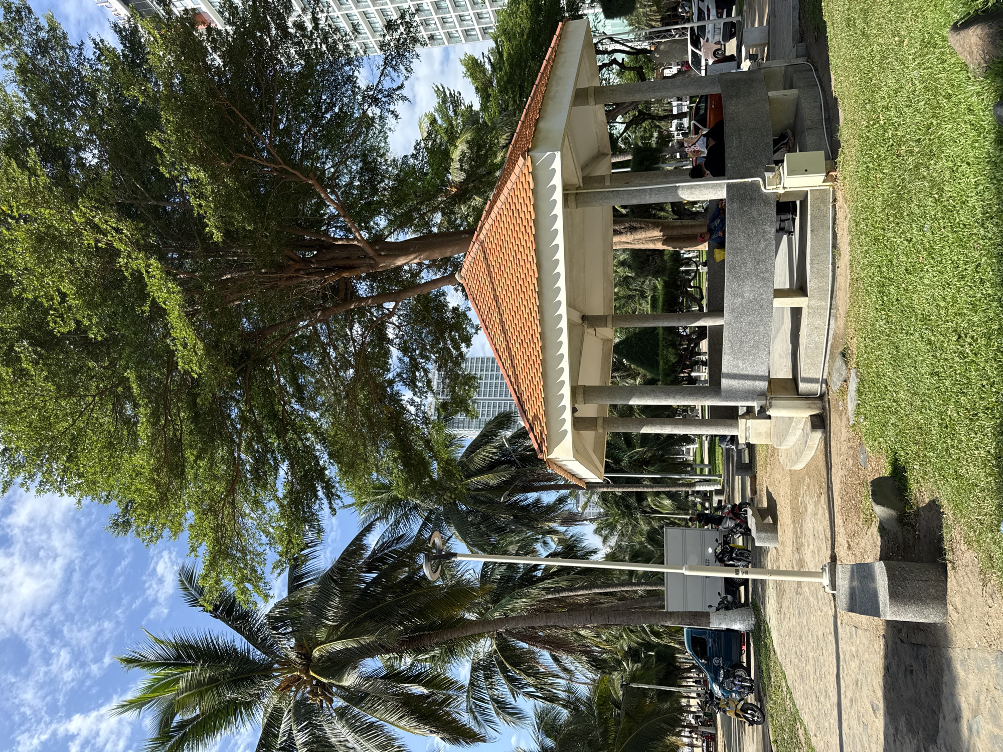 In Nha Trang, Vietnam, a sun-drenched scene unfolds under a bright blue sky dotted with scattered white clouds. The foreground is dominated by a circular, open-air pavilion with a terracotta-tiled roof and grey stone pillars. Inside, several individuals are resting, with some appearing to be asleep or relaxing. Around the pavilion, lush green grass covers the ground, interspersed with patches of dirt and stone pathways.
To the left of the pavilion, a paved walkway runs parallel to a grassy area. Parked along this walkway are several motorcycles and a light blue van. Palm trees, characteristic of a coastal environment, line the background, their fronds swaying gently. In the distance, modern high-rise buildings rise, suggesting an urban setting adjacent to a tropical landscape. The overall atmosphere is one of a peaceful public space, likely a park or recreational area near the coast.