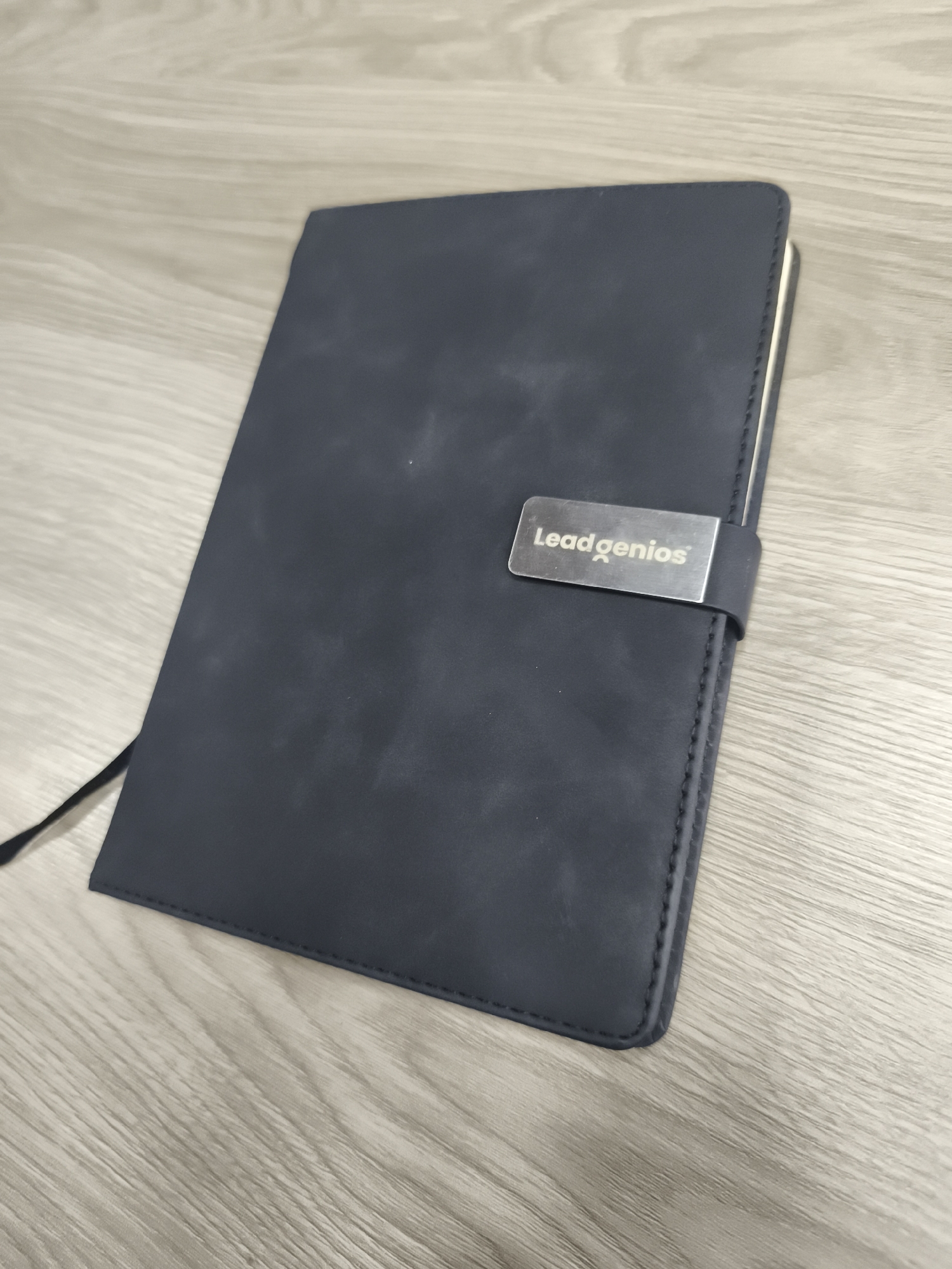 The image shows a closed, dark blue or gray notebook with a silver-colored clasp displaying the word "Leadgenios" in black text. The notebook is rectangular in shape with rounded corners and is sitting on a surface that appears to be a light-colored wood grain. A portion of a black elastic band is visible extending from the left side of the notebook. No people or other distinct objects are visible. The environment suggests an office or study setting, but there is no information to indicate that this photo was taken in Mexico City, Mexico.