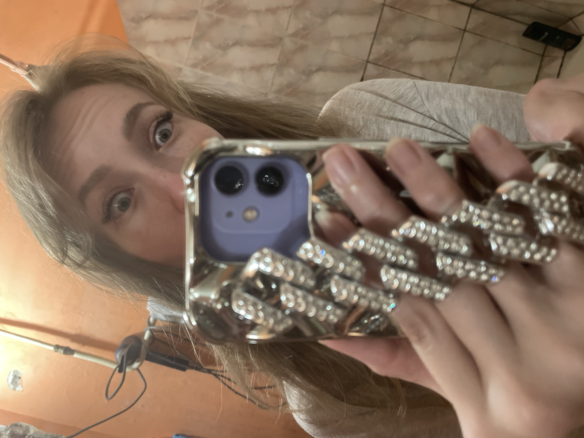 A person is holding a purple iPhone with a heavily jeweled case in front of their face, taking a selfie. The person has light skin and blonde hair. The background appears to be a room with peach-colored walls and tile. The image does not provide information to confirm the location in Perm, Russia.