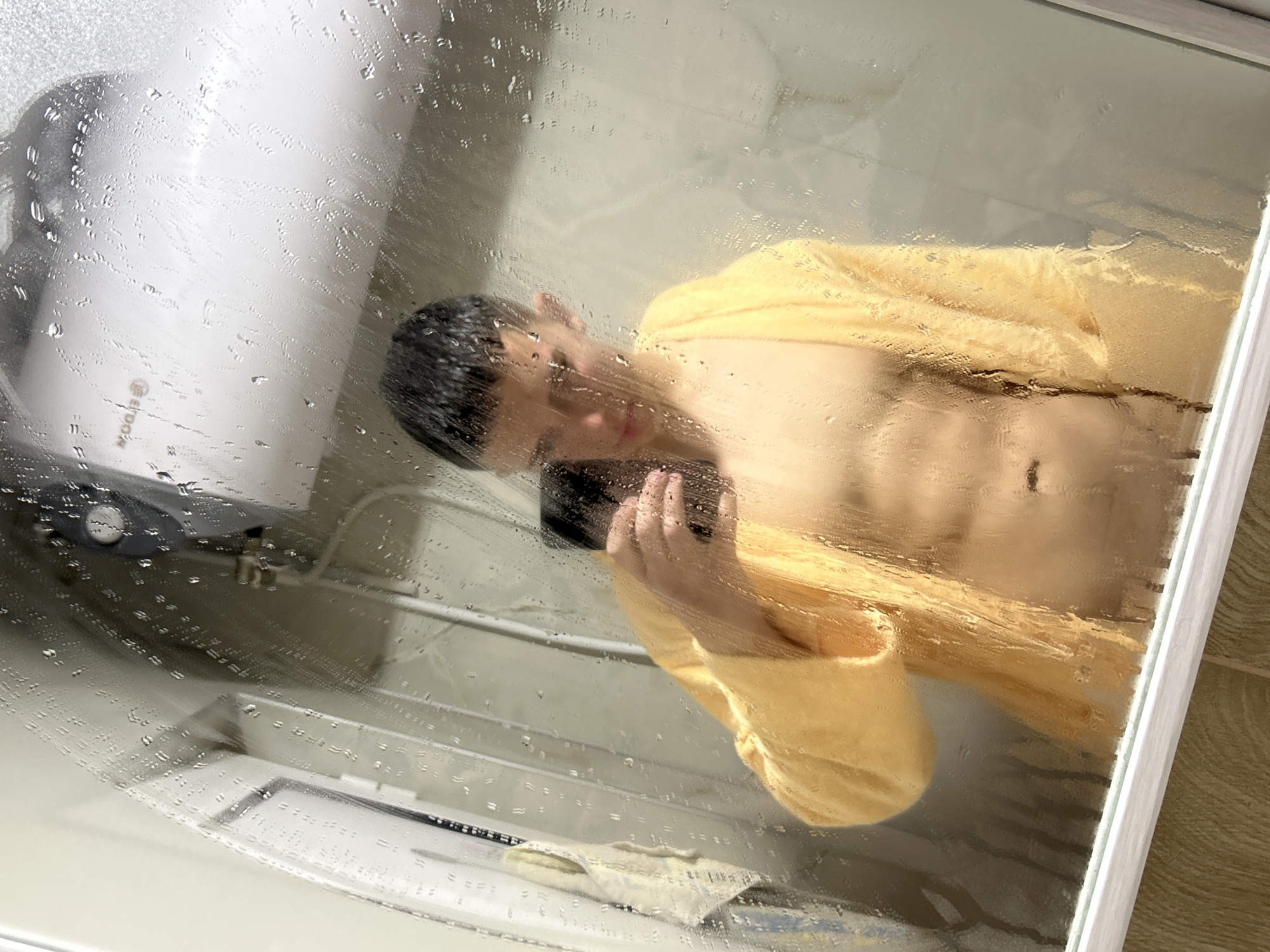 This indoor photograph, captured in Zlatograd, Bulgaria, depicts a person taking a mirror selfie in a steamy bathroom. The primary subject is a male-presenting individual with short dark hair, wearing a bright yellow bathrobe that is open to reveal his bare chest and abdomen. He is holding a black smartphone in his right hand, directing it towards the mirror to capture his reflection.
The mirror surface is prominently covered in a thick layer of condensation and numerous water droplets, which diffuse the light and slightly obscure the details of the reflection, creating a hazy, soft-focus effect. Through the mist, in the upper left of the reflection, a white water heater or boiler is visible, bearing the brand name "ELDOM" horizontally. Below the person's reflection, a portion of a bathroom sink or vanity counter with a folded light-colored cloth or towel can be seen. The steamy mirror suggests the person has recently had a hot shower or bath.