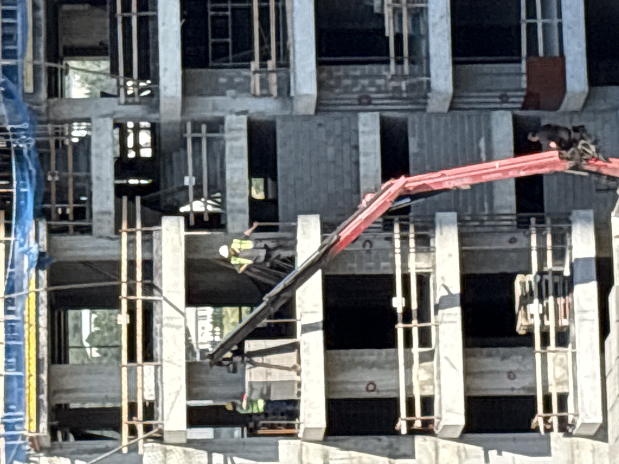 This is a medium-shot, eye-level, outdoor, daylight photograph of a multi-story building under construction. The building is constructed of concrete and cinder blocks, with wooden railings visible on several levels. A large red crane is positioned in the foreground, with its arm extending upwards and to the left, partially obscuring the building.
Two construction workers are visible on the second-floor level of the building. One worker, wearing a high-visibility vest and a hard hat, stands near a large grey object, possibly a load of building materials. The other worker, also wearing a high-visibility vest and a hard hat, is further to the right, holding onto some dark material. Another individual, dressed in dark clothing, is seated in the basket of the crane, near the lower portion of its arm.
The background shows more of the unfinished building, with open window frames and structural beams. Some greenery and parts of other buildings are visible through the open spaces. The overall impression is of an active construction site. This scene is located in Tbilisi, Georgia.