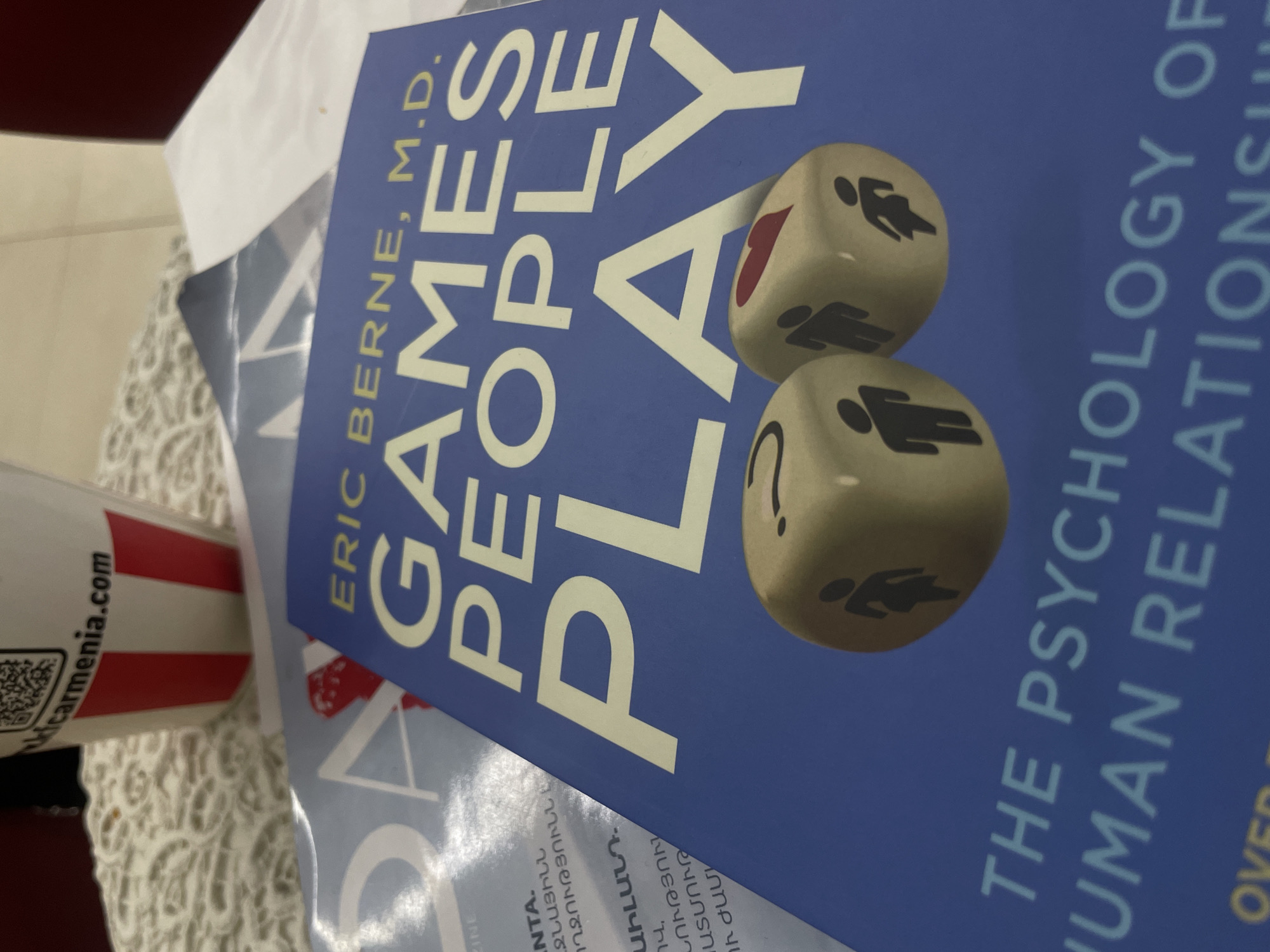 An angled, close-up shot features a copy of the book "GAMES PEOPLE PLAY" by Eric Berne, M.D., resting on a surface. The book has a blue cover with large, white sans-serif letters for the title and author's name. Part of the subtitle, "THE PSYCHOLOGY OF HUMAN RELATIONSHI", is also visible. The cover art includes two light-colored dice, one showing a red heart and a male stick figure, and the other displaying a question mark and a female stick figure.
Partially visible next to the book is a red and white striped cup with the website "kicarmenia.com" printed on it, alongside a QR code. Beneath the main book, hints of other documents or books are visible, displaying text in Armenian, including words like "ՋՆԱՅԻՆՆ", "ՈՂՋՈՒԹՅՈՒՆՆ Է", "ԱԻԼԱՆԴ.", and "ՆՈՒԹՅՈՒՆ". The scene is likely indoors, possibly on a table, in Yerevan, Armenia. No specific activity is occurring; it is a still life composition. The lighting appears soft and even.