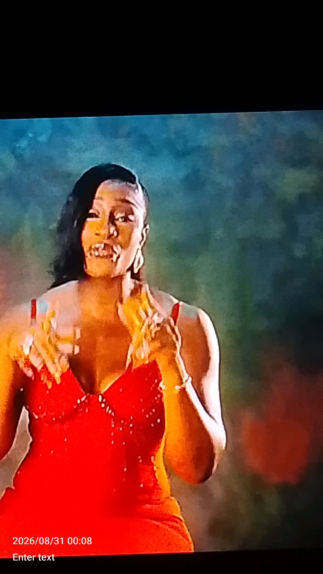 The image shows a woman wearing a red dress. She has dark hair and is wearing large earrings. She appears to be in motion, with her hands raised. The background is a blurry mix of green and brown. There is a timestamp in the lower left corner of the image that reads "2026/08/31 00:08". There is also text that reads "Enter text". The image quality is poor, with a lot of digital noise and compression artifacts, making it difficult to discern fine details. There is no discernible information about the location in Lokoja, Nigeria, or any specific activities or interactions.