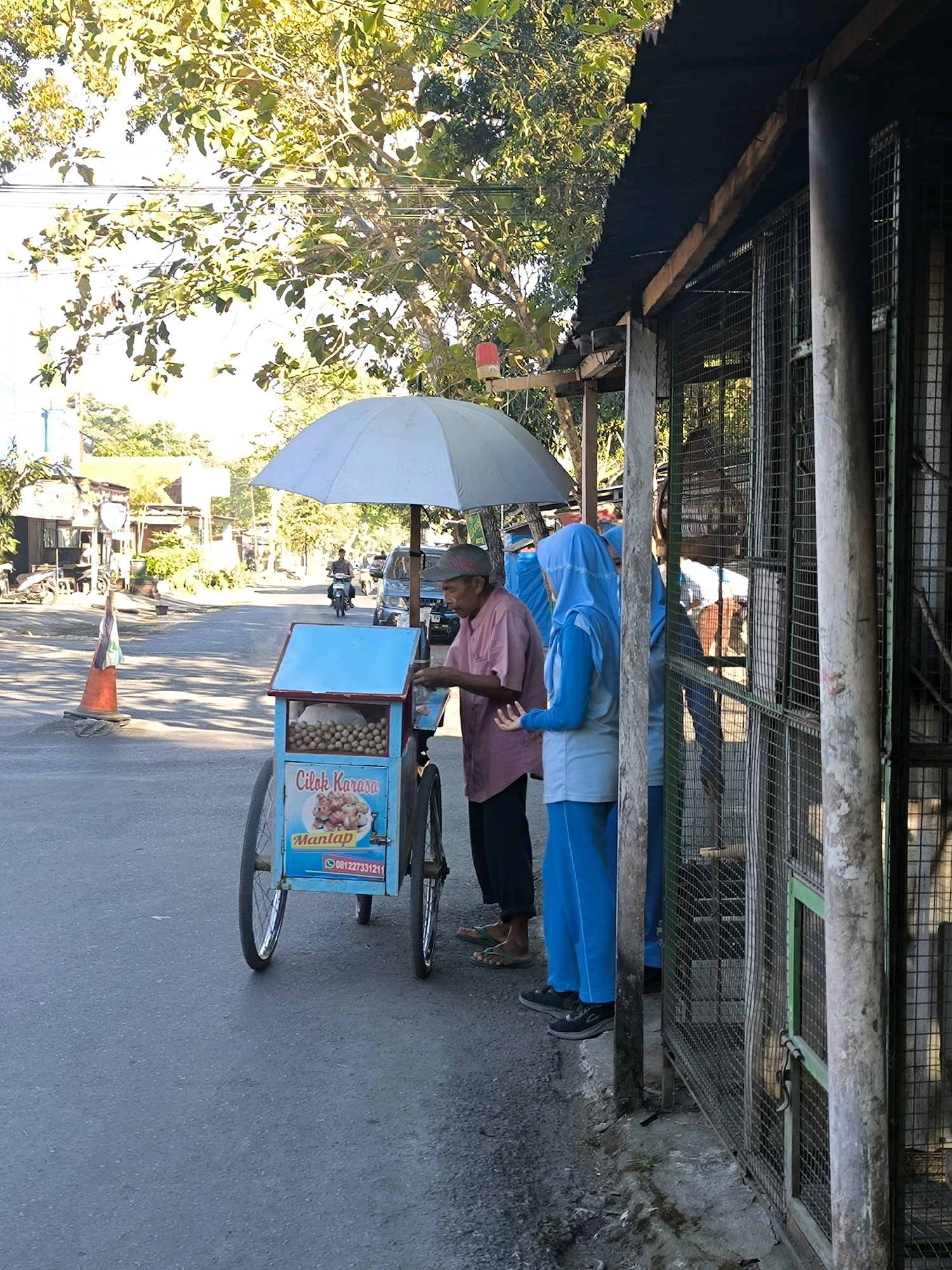 In Karanganyar, Indonesia, an elderly man stands beside a blue food cart, an umbrella overhead providing shade. The cart is filled with small, round food items, possibly meatballs, and displays text including "Cilok Karasa" and "Mantap," along with a phone number. Two women in blue hijabs stand to the right of the cart, near a structure with a wire mesh fence. The scene takes place on a paved street, with trees and buildings in the background. A motorcycle is visible in the distance on the road.