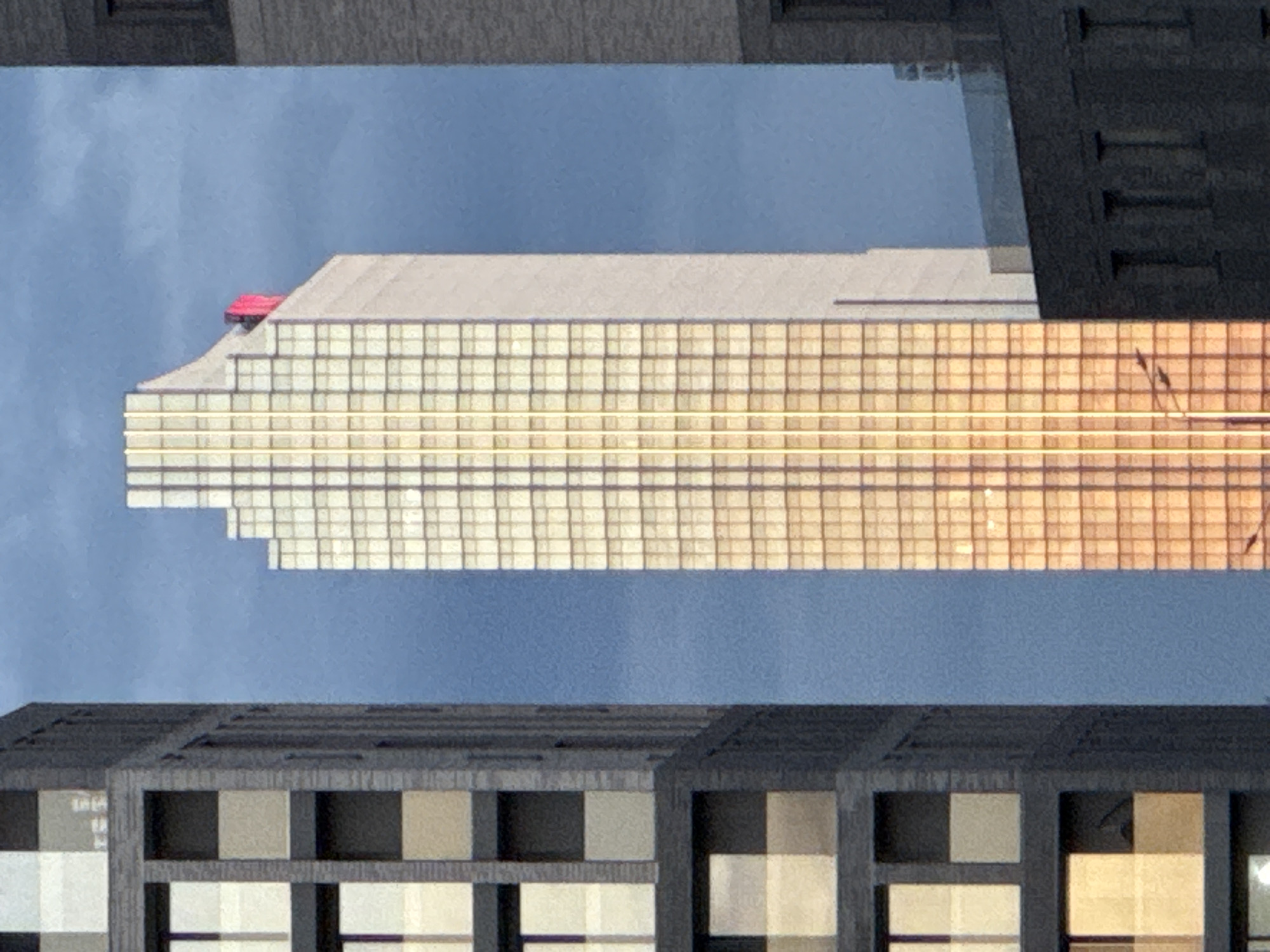 A vertically oriented photograph captures a striking urban scene, likely within Moscow, Russia, showcasing a modern skyscraper reflected in the glass facade of another building. The central subject is a tall building with a distinctive stepped and sloped roofline, predominantly light-colored at its upper sections. Its main facade, which appears golden due to sun reflections, is composed of a dense, rectilinear grid of glass panels.
The most notable and unusual detail is a bright red car, clearly identifiable as a Ferrari, positioned on the sloped upper section of the reflected skyscraper's roof. This car stands out against the light building material and the clear blue sky.
The central reflected building is framed by two other buildings, visible at the top and bottom of the image. The building at the top is dark grey with subtle texture, while the one at the bottom features a facade with large, square window-like sections in shades of grey and beige. A clear, uniform light blue sky is visible between these structures and the reflected skyscraper.
The golden hue on the reflected facade suggests that the photograph was taken during sunrise or sunset, with direct sunlight illuminating the building being reflected. There are no visible people or discernible text in the image. The scene highlights architectural scale, reflections, and an unexpected artistic or commercial placement of a luxury vehicle atop a skyscraper.