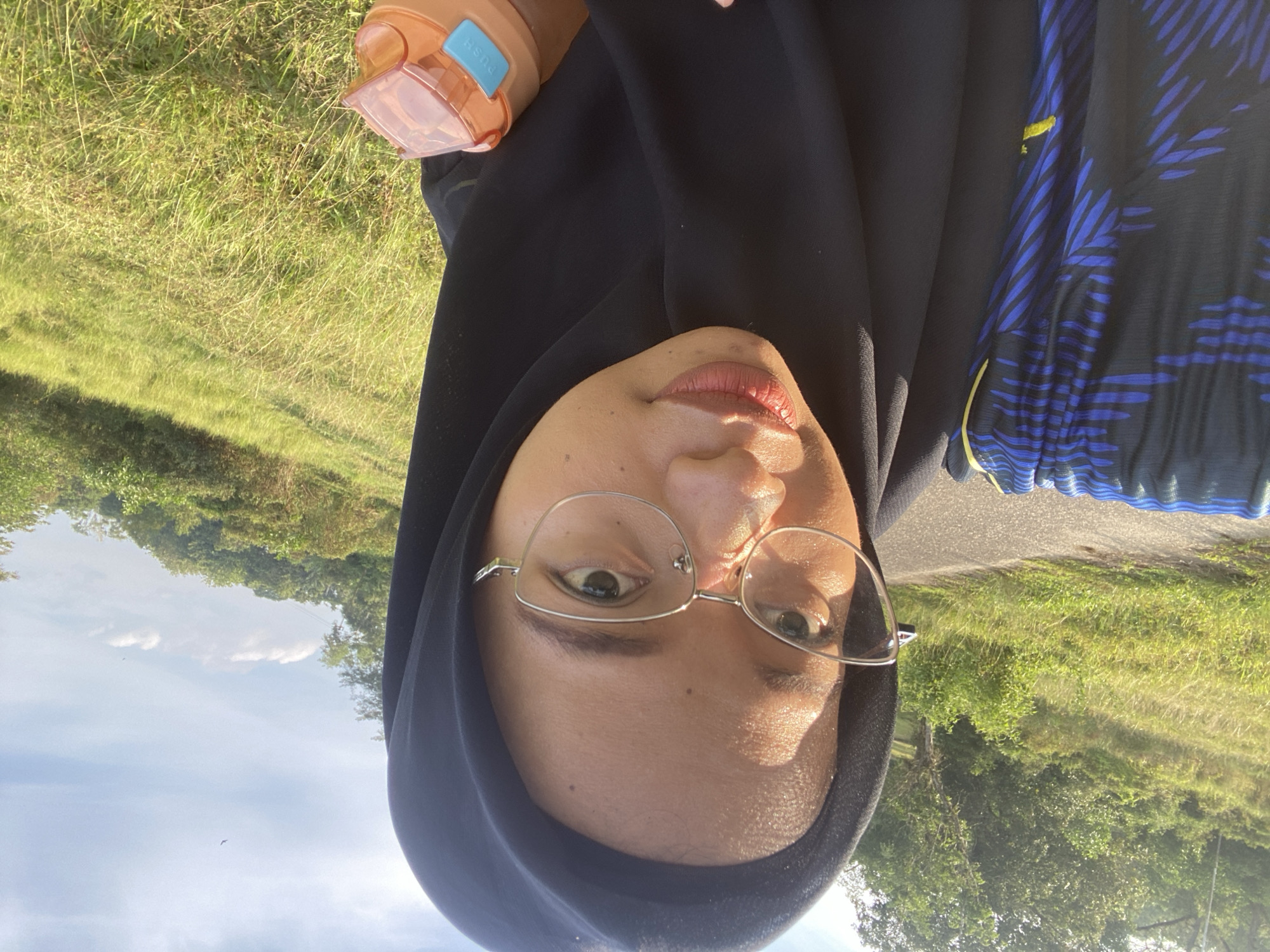 A young woman wearing a black hijab and eyeglasses is taking a selfie outdoors in a grassy, hilly area. She is wearing a black and blue patterned shirt. She holds an orange and blue water bottle in her right hand. The background shows a road, green trees, and a bright blue sky with some clouds. The location is the Gudang Batu area in Indonesia.