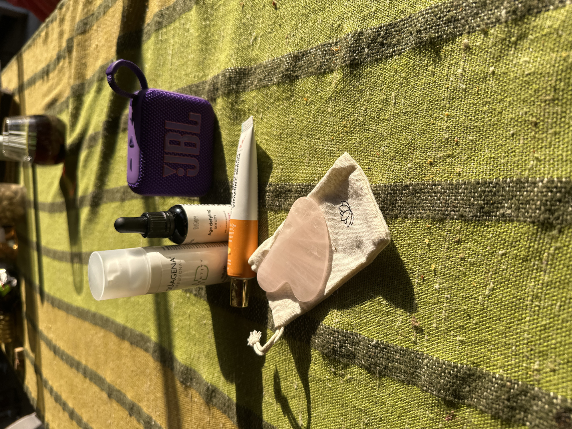 A top-down shot reveals a JBL Bluetooth speaker, skincare products including serums and creams, and a rose quartz Gua Sha tool nestled in a small drawstring bag with a lotus flower design, arranged on a green and tan striped woven blanket. The blanket is laid out in sunlight, likely outdoors, possibly in a city (Pliska, Bulgaria is mentioned, suggesting the location is within that city).