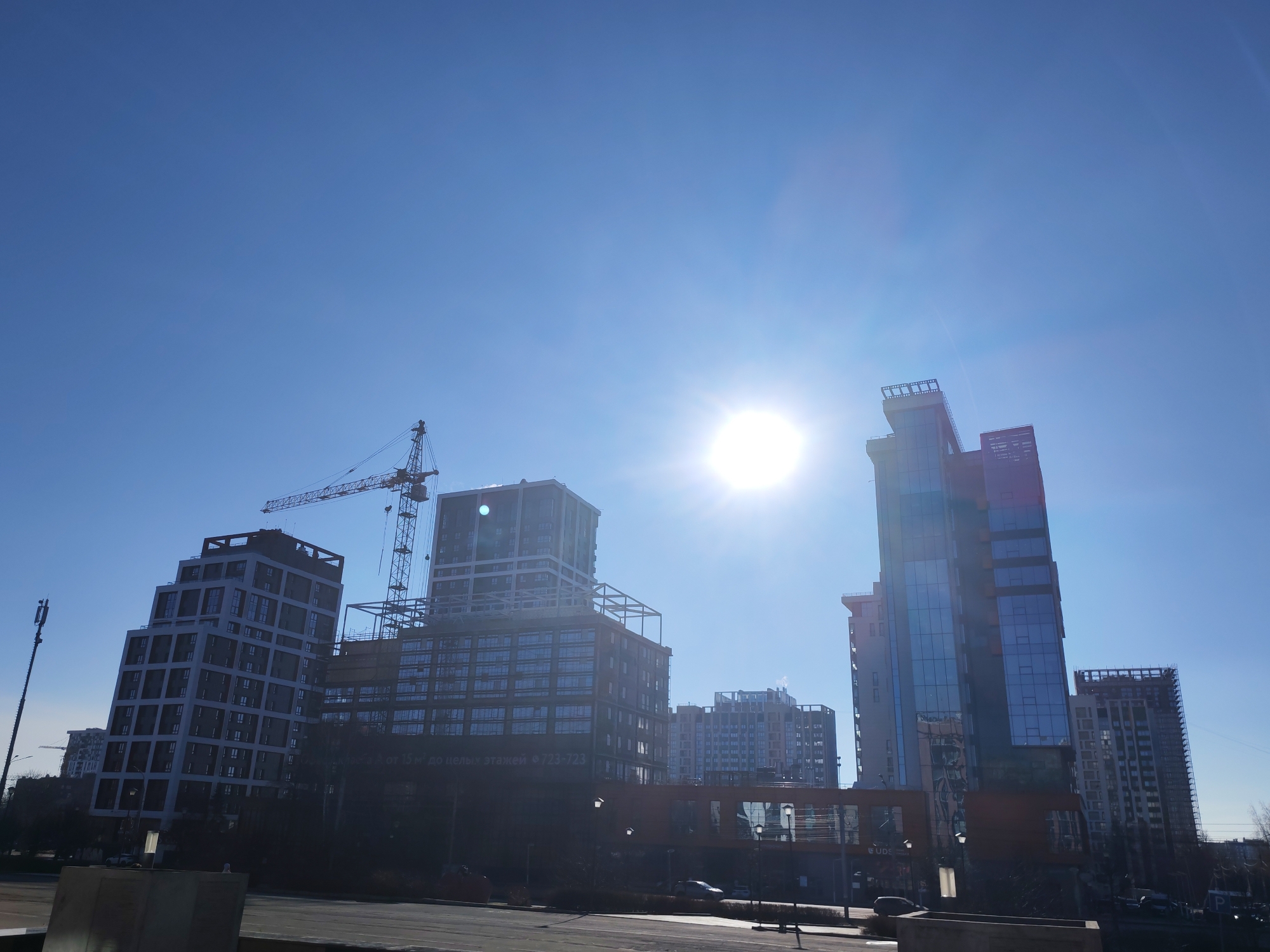 The image depicts a cityscape in Izhevsk, Russia, dominated by several high-rise buildings, some still under construction. A construction crane is visible near one of the buildings. The sky is clear and bright blue, with the sun positioned centrally, creating a strong glare. The buildings vary in design, some with traditional rectangular structures and others with more modern, glass-clad facades. A roadway is in the foreground, and a few cars are visible on the road. Streetlights are also present. The overall scene suggests an urban environment with ongoing construction and development.