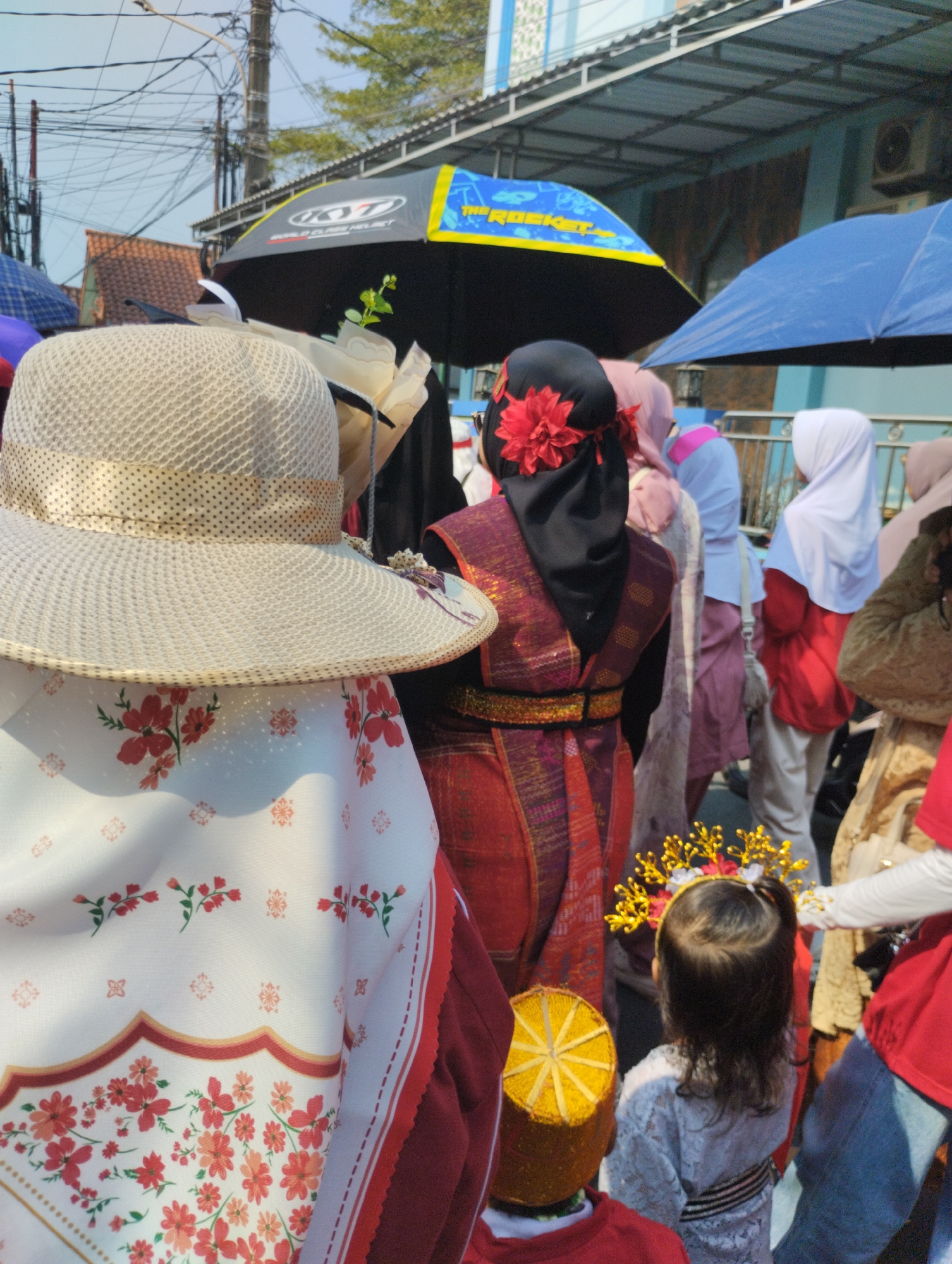 The image depicts a street scene, likely in Bekasi, Indonesia, on a sunny day, with several individuals and umbrellas visible. In the foreground, a person wearing a wide-brimmed, woven hat with a patterned ribbon and a white garment adorned with red floral motifs is positioned centrally. To their right, another person in a black headscarf with a prominent red flower accessory and a traditional red and gold patterned outfit is also in focus. Behind them, a group of people are gathered, some holding umbrellas, including a large black umbrella with "KYT" branding and a blue umbrella. The background reveals buildings with a blue facade and awnings, as well as overhead power lines, suggesting an urban setting. A young child wearing a light-colored, patterned outfit and a golden headdress is visible in the lower right portion of the image. The overall atmosphere suggests a public gathering or event.
