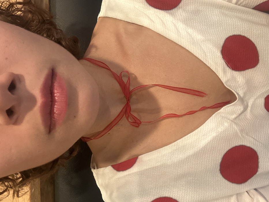 A close-up, slightly angled shot shows a person's face and upper torso. Prominent features include full, glossy lips, nose, and curly hair. A red ribbon is tied around the neck, resembling a delicate necklace. The person wears a white top adorned with large, evenly spaced red polka dots. The skin tone appears fair. The photo is taken from a low perspective.