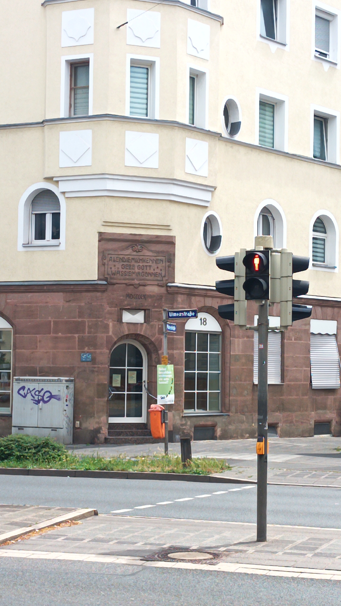 This image shows a street scene in Nuremberg, Germany, dominated by a building with light yellow upper floors and a red brick lower facade. A traffic light with a red pedestrian signal is prominently in the foreground. The building features several windows, some with arched tops and others rectangular. Decorative white squares are visible on the upper facade. Above the red brick section, an inscription is carved into the stone.
The scene appears to be on a street corner, with a street sign reading "Ulmerstraße" and the number "18" visible on a pole. An orange trash can is mounted on a pole near the building's entrance. Graffiti is present on a metal utility box to the left of the entrance. Grass and some greenery are visible along the sidewalk. The pavement and road are also visible, with a manhole cover in the foreground. The overall impression is of a typical urban environment.
The red pedestrian signal indicates that it is currently not safe for pedestrians to cross the street. The time of day is not clearly discernible, but the lighting suggests it could be daytime. No people are clearly visible in the image.
The inscription on the building's facade reads: "ALLEN DIE MIHMKENNEN GEBE GOTT, WAS SIE MIR GANNEN. MDCCCCX".