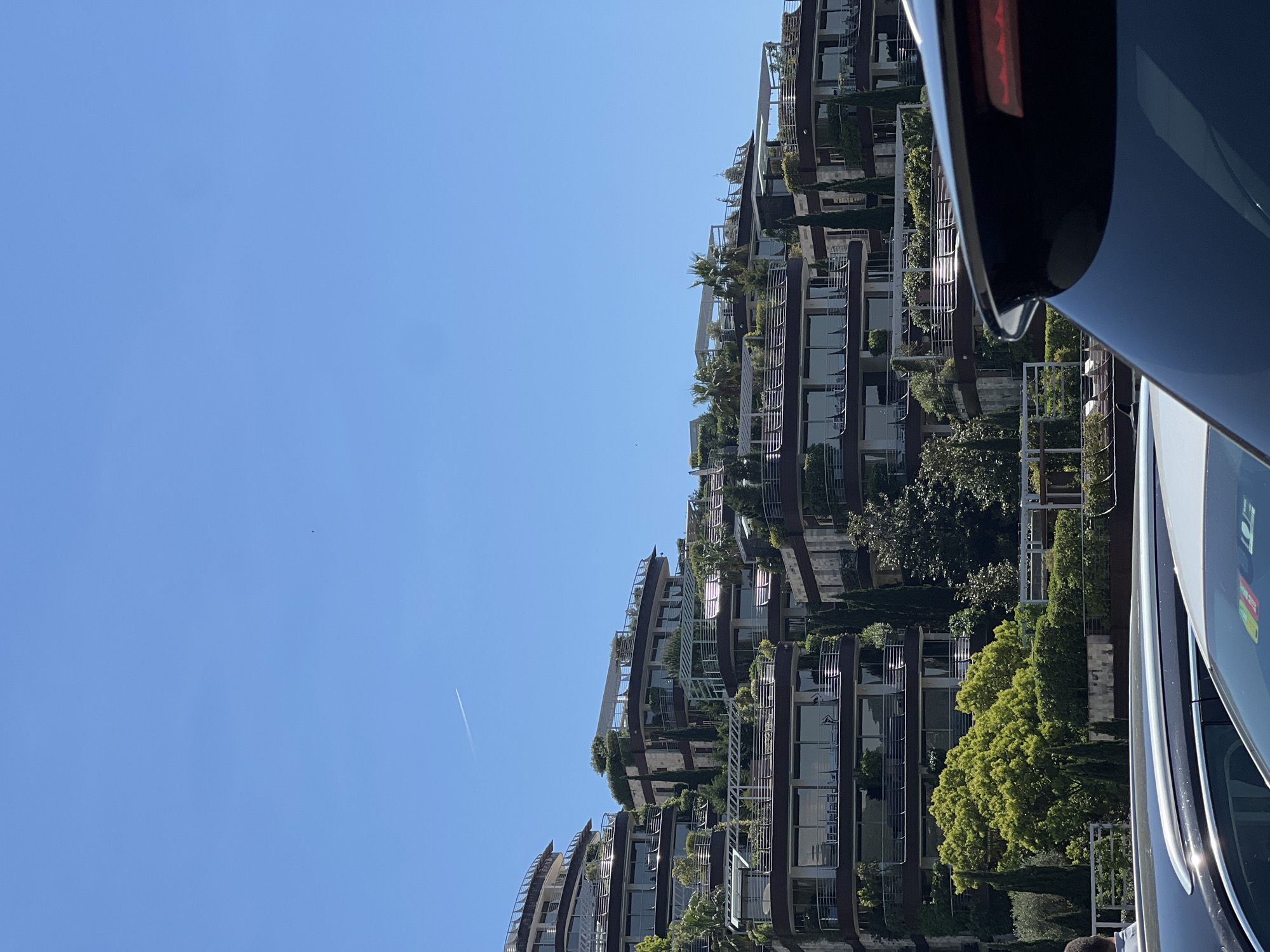 The image captures a modern, multi-story building featuring green architecture, set against a clear blue sky, with a dark vehicle partially visible in the foreground. The building, which dominates the middle and left side of the frame, boasts numerous balconies and terraces extensively integrated with lush vegetation, including small trees, shrubs, and climbing plants, creating a verdant, cascading effect. Its facade appears to be constructed with dark paneling, glass windows, and light-colored metal railings.
In the expansive, bright blue sky above the building, a distinct white contrail from an airplane stretches diagonally across the upper left. A tiny dark speck, possibly a bird, is also visible further left in the sky.
On the immediate right side of the foreground, a section of a dark, reflective vehicle, possibly a car or a bus, occupies the edge of the frame. A red light, indicative of a taillight, is visible on its upper right side, along with a silver or white trim line. Some faint, unreadable text or graphics are visible on the vehicle's dark surface.
The scene depicts a bright, sunny daytime with clear weather. The modern architectural style, emphasizing natural integration, is prominent. No specific human activity is taking place. The location of the scene is Budva, Montenegro.