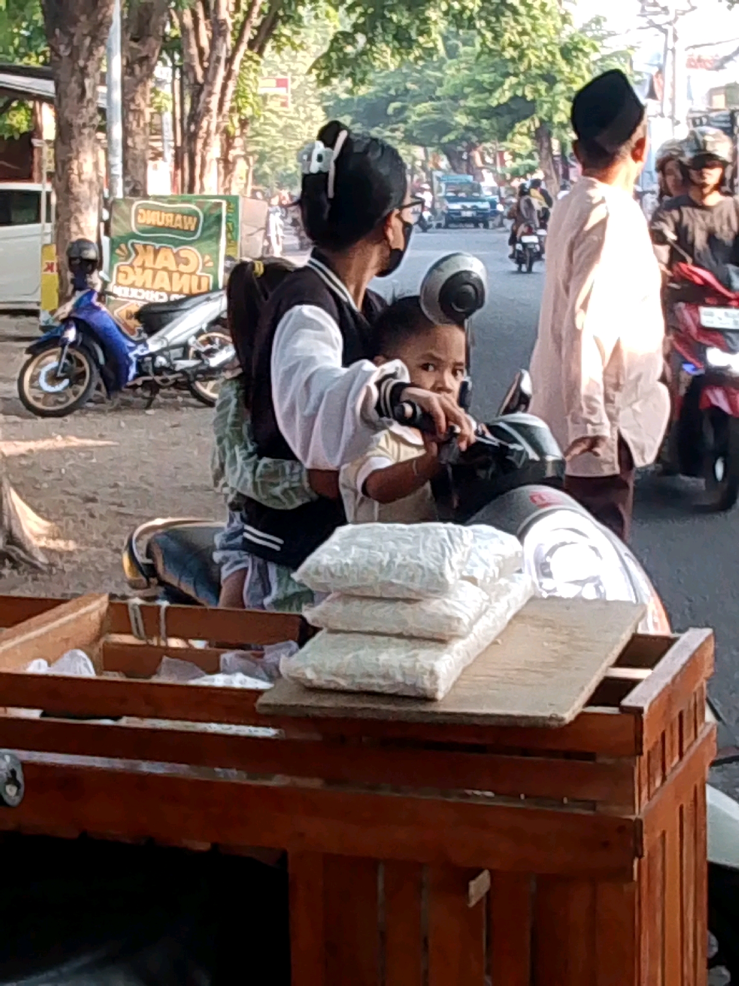 This image depicts a street scene in Probolinggo, Indonesia. In the foreground, a wooden cart is filled with items, possibly merchandise. On top of the cart, there are several white packages. Behind the cart, a child is seated on a motorcycle, looking towards the camera. The child is wearing a light-colored shirt. A woman, presumably the child's mother, is seated behind the child on the motorcycle, holding onto the child. She is wearing a dark jacket over a white garment and has her hair pulled back. To the right of the motorcycle, a man in a light-colored long-sleeved shirt and a black cap is visible. In the background, other motorcycles and people are present, indicating a busy street. A sign with text in Indonesian can be seen on a green background to the left of the motorcycle. The environment appears to be an urban setting with trees lining the street.